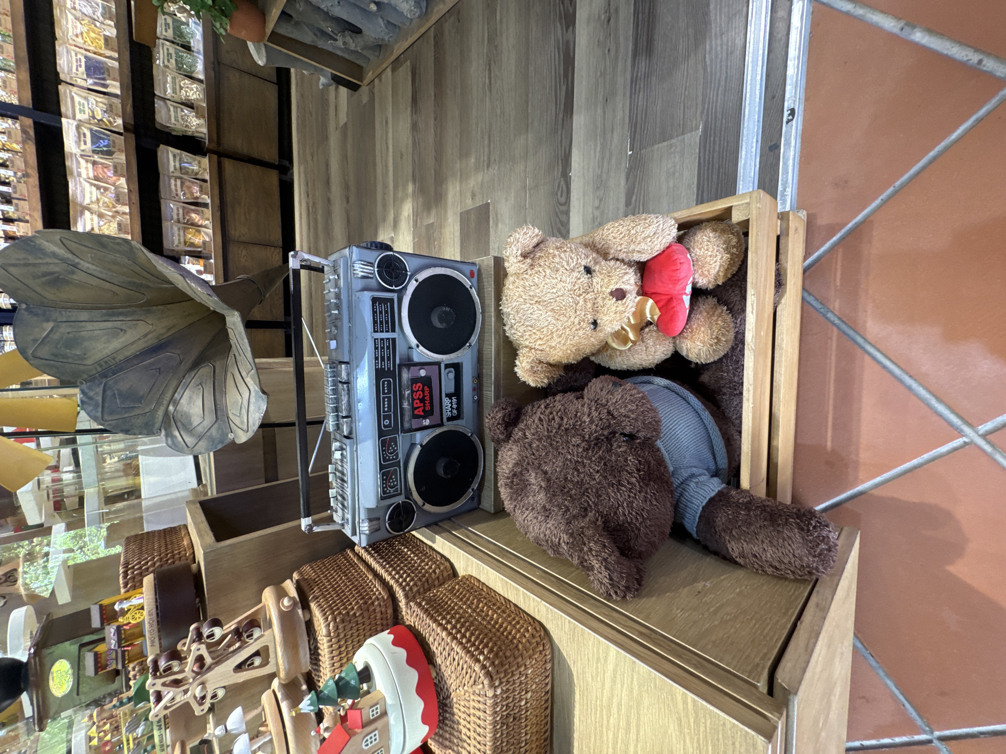 The image shows a display shelf in a store. In the top right corner, two teddy bears are crammed into a small wooden crate. One bear is light brown and holds a red heart. The other bear is darker brown and wears a grey shirt. To the left of the crate sits a grey boombox. The boombox is placed on top of several small wicker baskets. In the background, a collection of goods are on display. A gramophone sits at the front of the display. The location is likely a shop in Đà Nẵng, Vietnam.