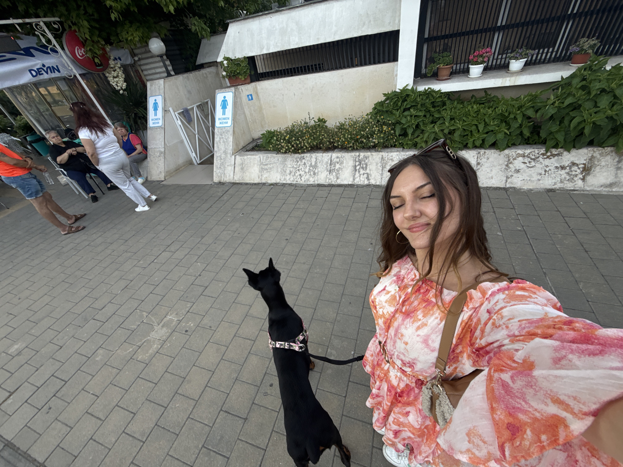 The image displays a detailed outdoor scene in Burgas, Bulgaria, captured from a selfie perspective. In the immediate foreground, to the right, a young woman with long, dark brown hair, wearing sunglasses on her head and a multi-colored (pink, orange, white) patterned dress with ruffled sleeves, is shown with closed eyes and a slight smile, taking the photograph. A thin brown strap from a shoulder bag is visible across her chest. Adjacent to her, a small to medium-sized black dog, wearing a multi-patterned harness and a black leash, stands facing away from the camera, looking towards the left side of the frame. Both the woman and the dog are standing on a surface composed of light grey rectangular paver blocks.
In the midground, to the left, several individuals are present beneath a white canopy structure with visible signage for "DEVIN" and "Coca-Cola." One person, seen from the back, wears an orange shirt and blue shorts, holding a water bottle. Two older women are seated at a table, one in a dark top and the other in a blue top. A fifth person, a woman with dark hair wearing a white top and light-colored pants, is walking away from the camera towards a building entrance.
The background features a light-colored concrete building. Prominently displayed on the building are two blue public restroom signs, each featuring a white female pictogram and the text "WOMEN / ЖЕНИ" (Women / Women in Bulgarian Cyrillic). A white metal gate structure is visible near these signs. Below a section of the building, a planted area contains green foliage and small yellow flowers. Higher up, on a narrow ledge, several potted plants with pink and purple flowers are arranged. The setting is indicative of a public or commercial area, possibly a park or promenade, within the city of Burgas, Bulgaria.