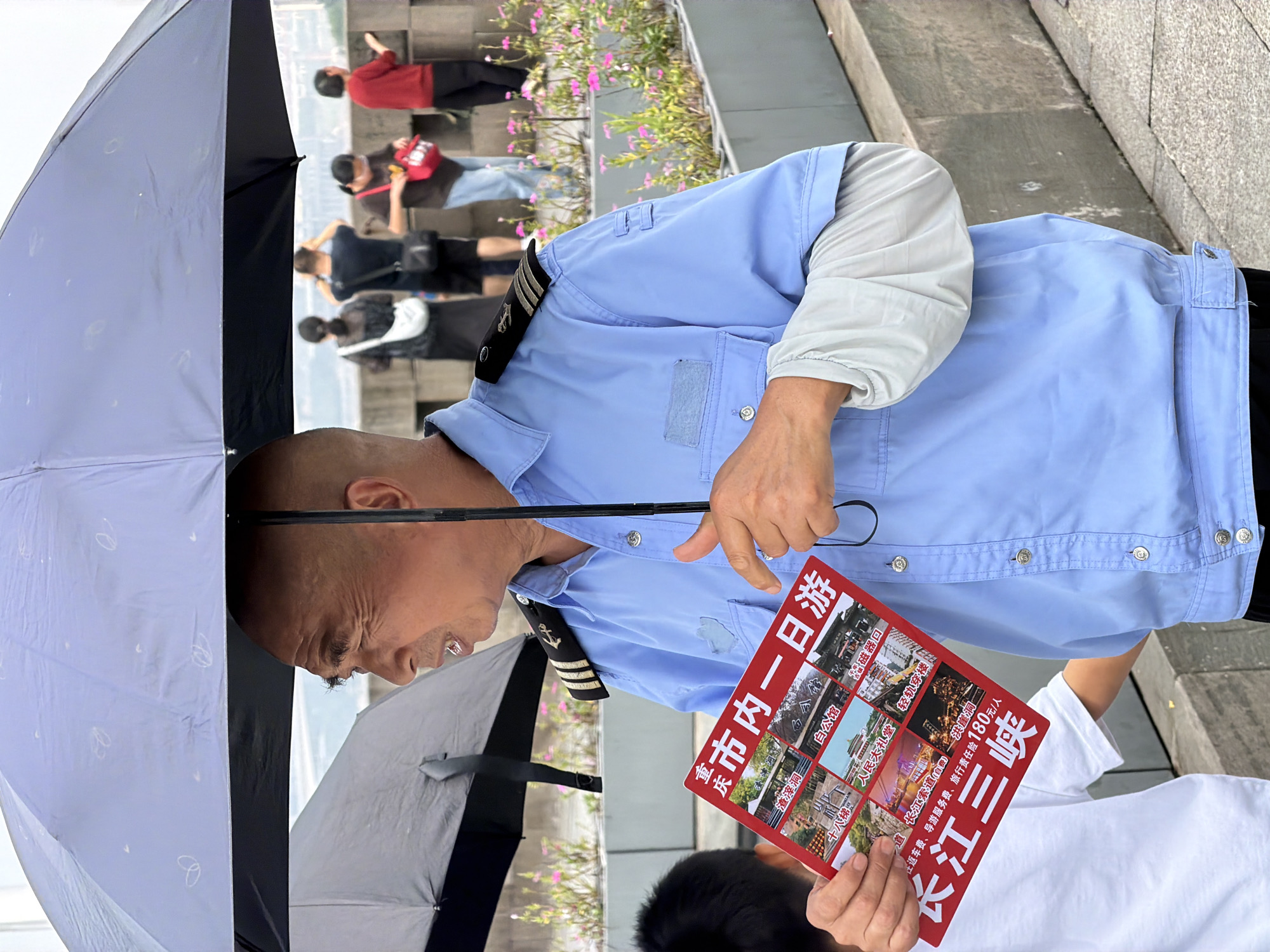 An East Asian man, appearing to be in his 60s, stands partially obscured by a dark umbrella, holding a red brochure. He wears a light blue collared shirt with epaulets indicating rank, suggesting he might be a uniformed official or tour guide. The brochure features multiple images of tourist attractions and text in Chinese characters, with "长江三峡" (Yangtze Three Gorges) visible at the bottom. The man is looking to his left and speaking, with his right hand gesturing towards the brochure. A younger East Asian person in a white t-shirt is partially visible to his left. In the background, several other individuals, likely tourists, stand near a low wall with planters containing pink flowers. The setting appears to be an outdoor public area, possibly a waterfront promenade in Yuzhong District, Chongqing, China, given the "Yangtze Three Gorges" reference and the urban landscape visible in the distance. The overall atmosphere is casual, with overcast conditions suggested by the umbrella.