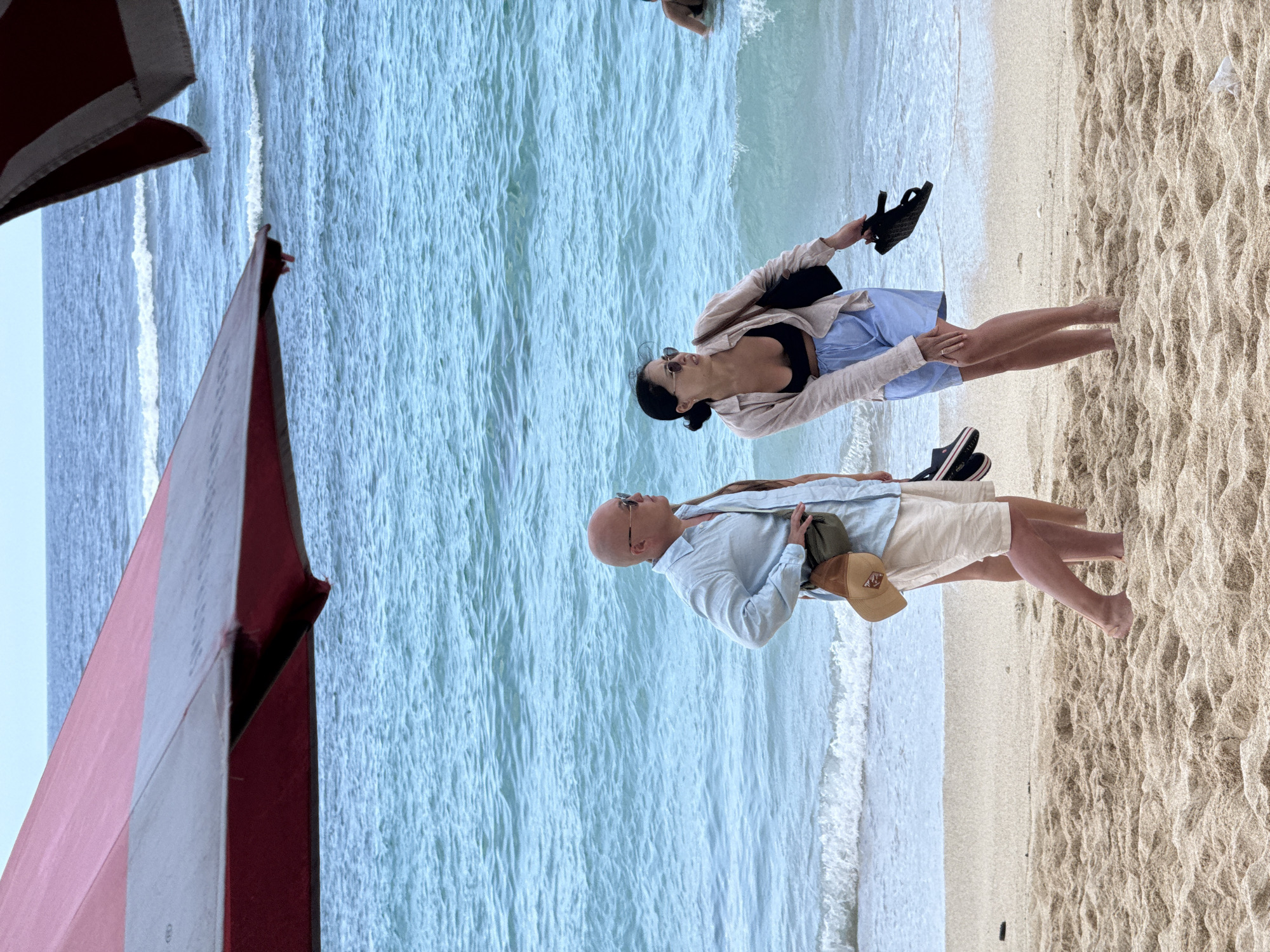 The image shows a man and a woman standing on a beach in Kuta Selatan, Indonesia. The man is bald and is wearing a light blue shirt, tan shorts, and a baseball cap. The woman is wearing a black swimsuit top, a light-colored shirt, and a blue skirt. She is also wearing sunglasses. The man is facing toward the ocean, and the woman is facing away from the ocean. The waves are gently crashing on the shore. In the foreground, there is part of a structure with red and white stripes.