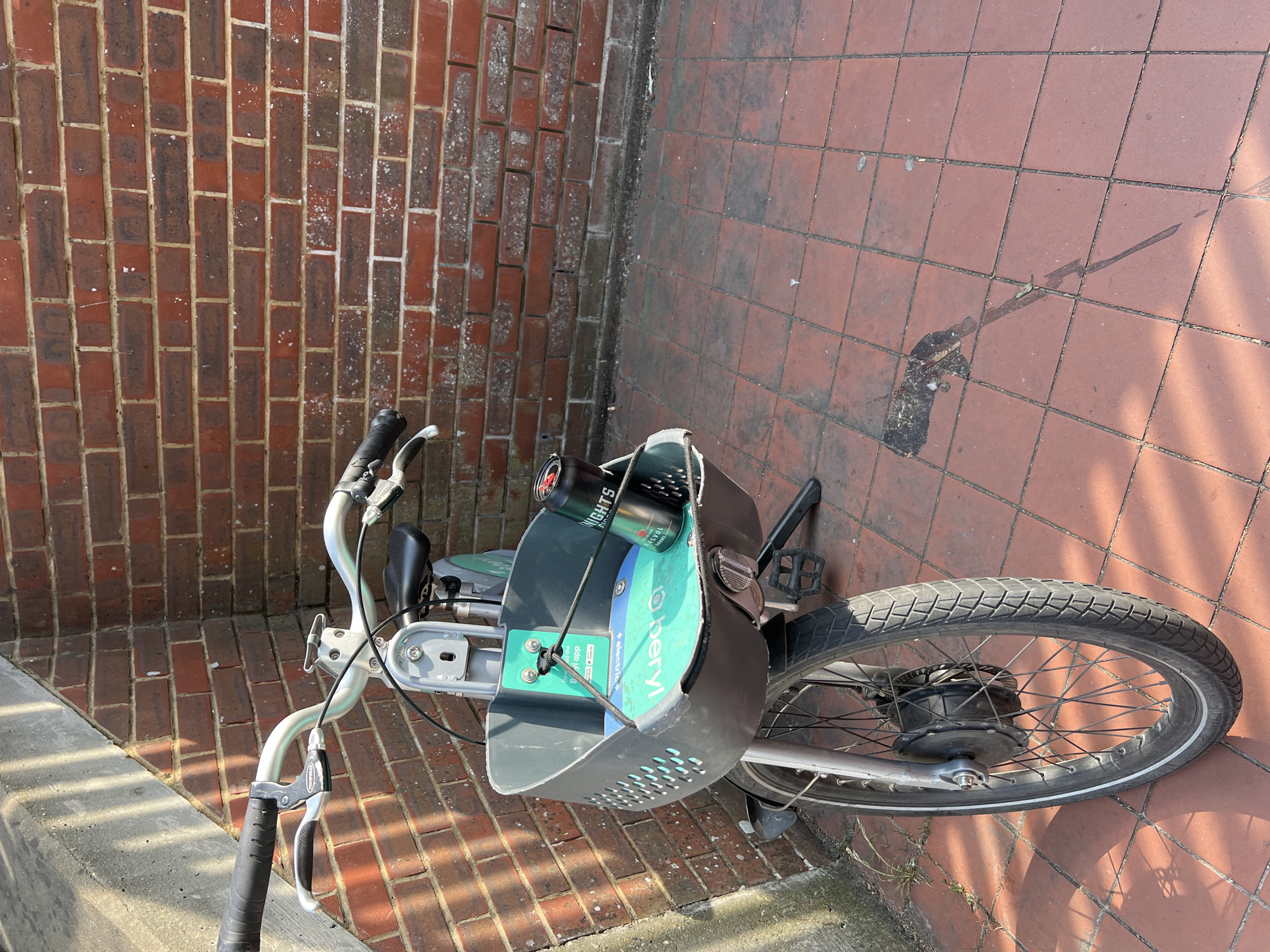 An abandoned electric bicycle with a black can of "Knights" beverage resting in its front basket is parked against a red brick wall, next to a concrete structure with sunlight casting diagonal shadows. The bicycle has a grey plastic basket, a black seat, and a front wheel with a motor hub. The ground is paved with red tiles. A dark, wet stain is visible on the tiled ground. The scene appears to be in an outdoor public space in Guildford, United Kingdom, based on the "o-Bikes" branding visible on the bike, which was a former bike-sharing company that operated in the UK.