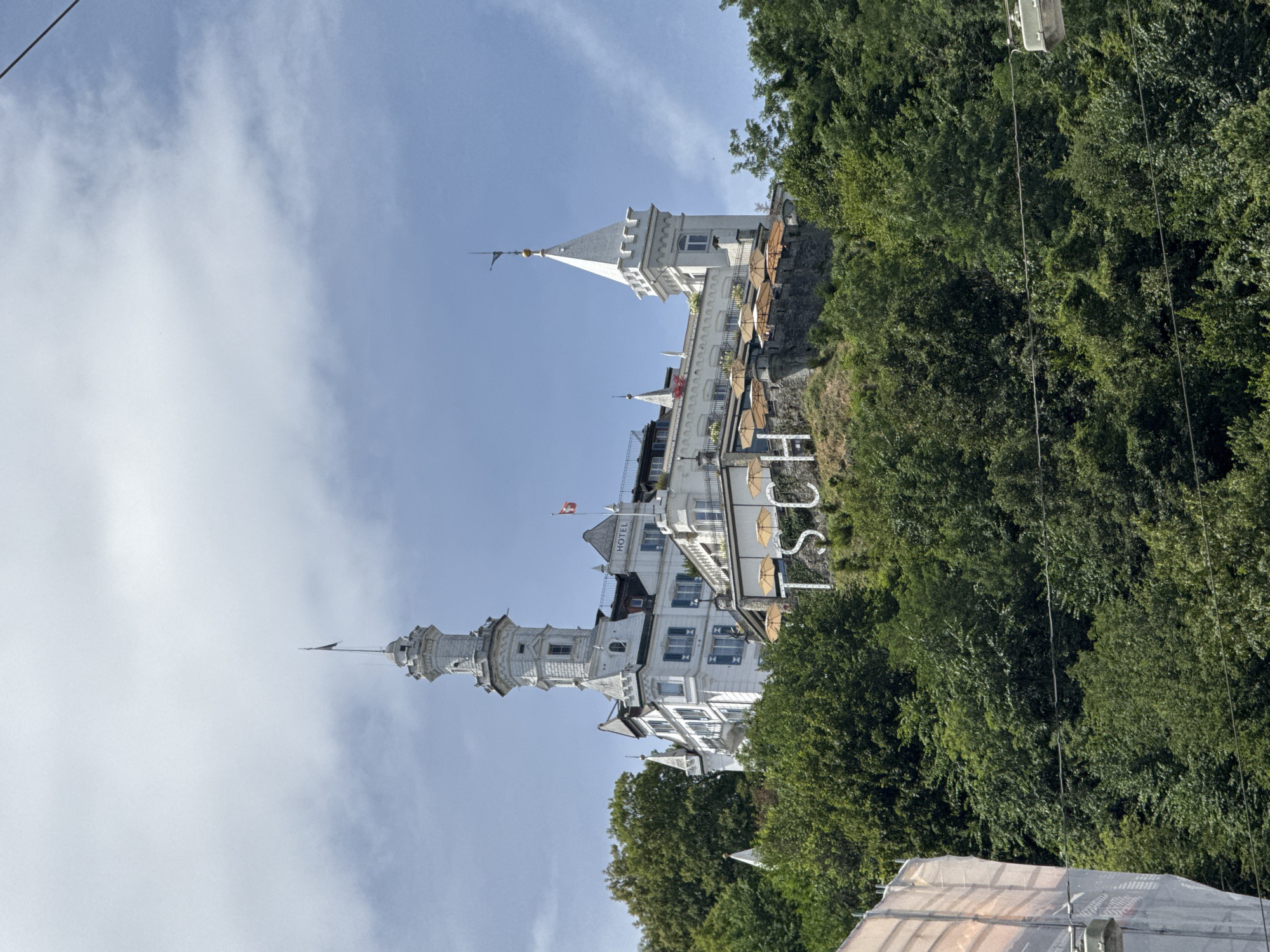The image shows the historic Victoria Jungfrau Grand Hotel & Spa, a prominent landmark in Interlaken, Switzerland, not Lucerne, situated atop a hill overlooking lush green trees. The grand white hotel features distinctive architecture with multiple towers, turrets, and ornate detailing. A large white sign spelling "HOTEL" is visible near the top of the building. Below the hotel, large stone letters forming "TSC" are embedded in the hillside, possibly part of the hotel's branding or a historical marker. Outdoor seating with umbrellas is set up on a terrace overlooking the town. The sky is partly cloudy with a clear blue sky visible. In the foreground, the edge of a building covered in scaffolding and netting is partially visible on the left, and power lines traverse the lower portion of the image. The overall impression is a majestic hotel in a scenic, verdant setting.
