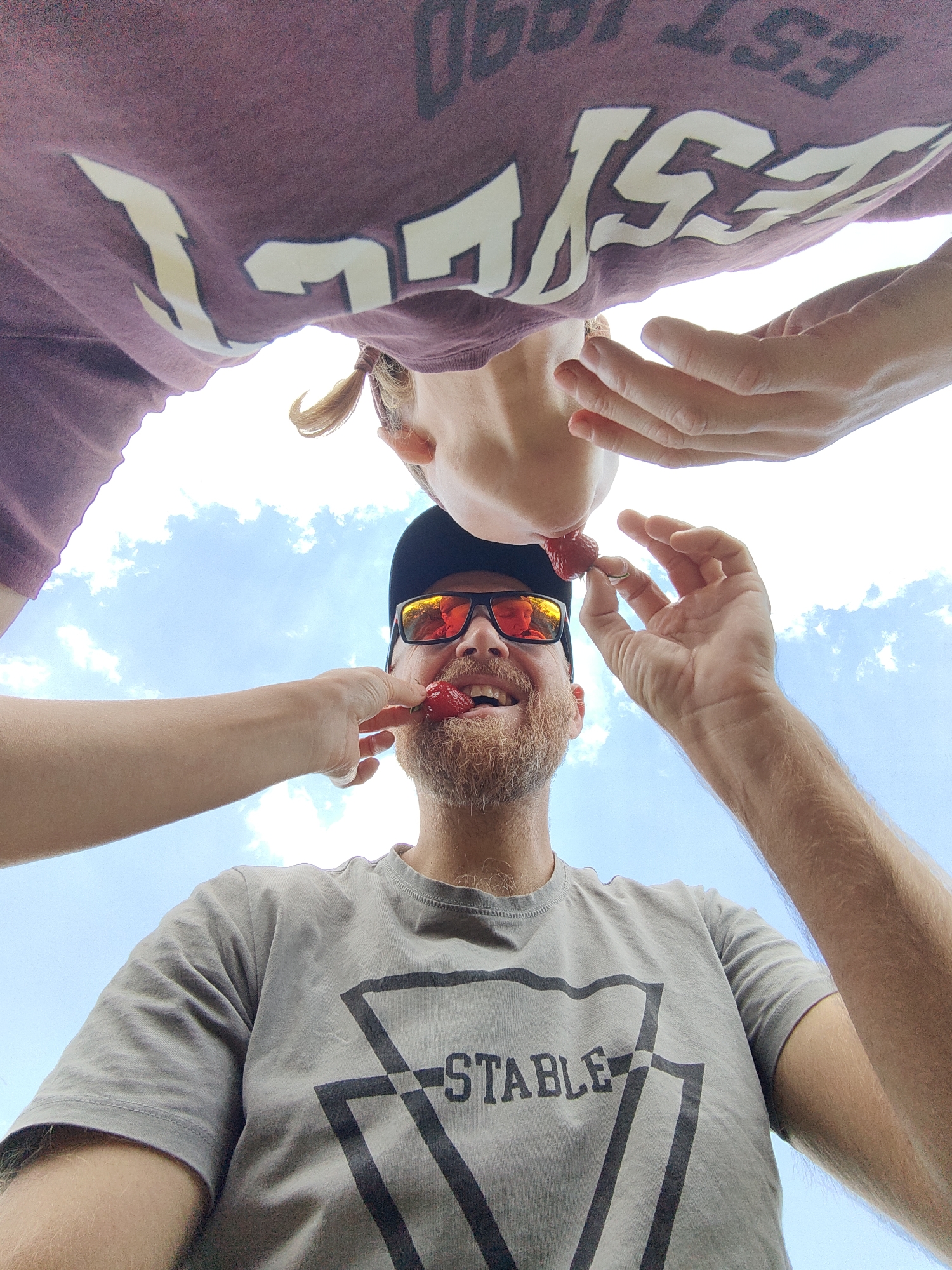 An extreme low-angle, wide-shot photograph captures a couple sharing strawberries against a backdrop of a bright blue sky with scattered white clouds. The perspective is from below, looking up at the subjects.
In the foreground, a man wearing a gray t-shirt with a black geometric logo and the word "STABLE" is positioned centrally. He has a beard and is wearing black sunglasses with orange mirrored lenses that reflect the sky and possibly the person taking the photo. He is smiling, with his mouth slightly open, and a strawberry is partially in his mouth, being held by his right hand. His left hand is extended upwards, holding another strawberry near his mouth.
Above the man, a woman is positioned, her face angled downwards. She is wearing a maroon t-shirt with a large white graphic that is partially obscured. Her blonde hair is tied back in a ponytail. She is holding a strawberry near the man's mouth with her left hand, and her right hand is also extended. It appears she is feeding the man a strawberry, and he is simultaneously feeding himself or the woman.
The environment is an outdoor setting, indicated by the clear sky and clouds. The overall scene suggests a playful and intimate moment between the couple. There are no visible buildings or specific urban or rural landmarks. The location is not identifiable from the visual information provided.