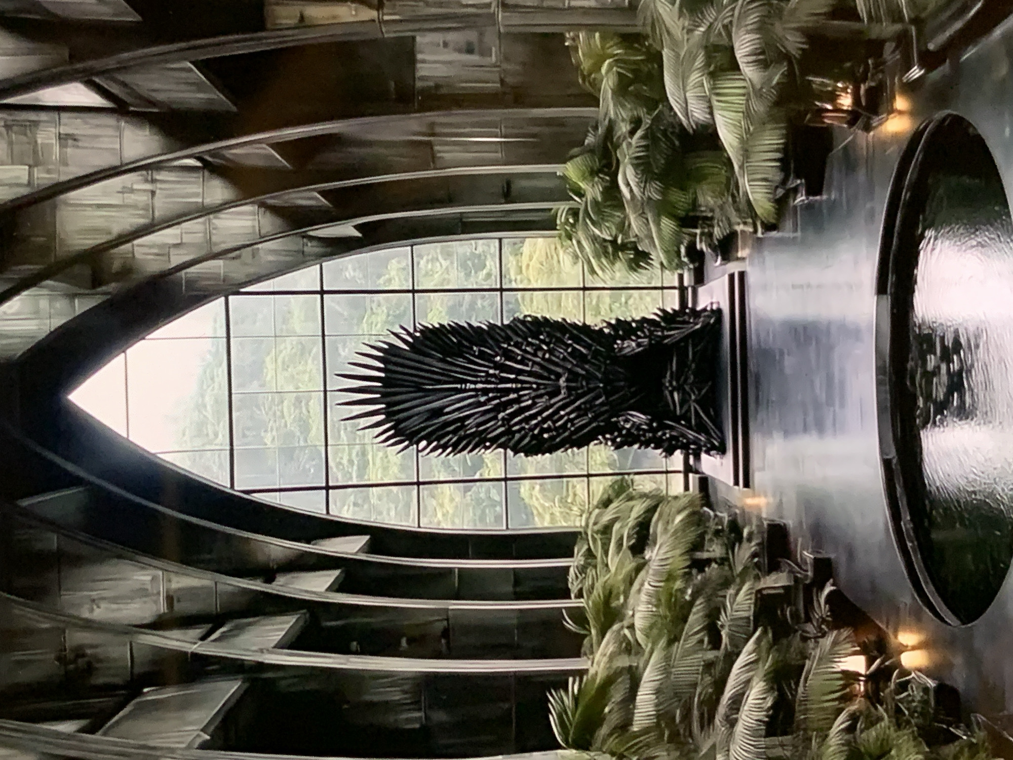The image depicts a grand, dimly lit interior space with a prominent, imposing throne at its center. The throne appears to be made of dark, metallic material, resembling swords or sharp points, and is situated in front of a large, arched window that looks out onto a lush, green landscape. Flanking the throne are rows of potted palm plants, their fronds creating a dense, verdant border. The floor in front of the throne is highly reflective, mirroring the surrounding architecture and greenery, and features a circular water feature. The architecture itself is characterized by dark, curved elements that suggest a Gothic or dramatic style. The overall ambiance is one of power, mystery, and a touch of the theatrical.
This image is NOT located in Stavropol, Russia.