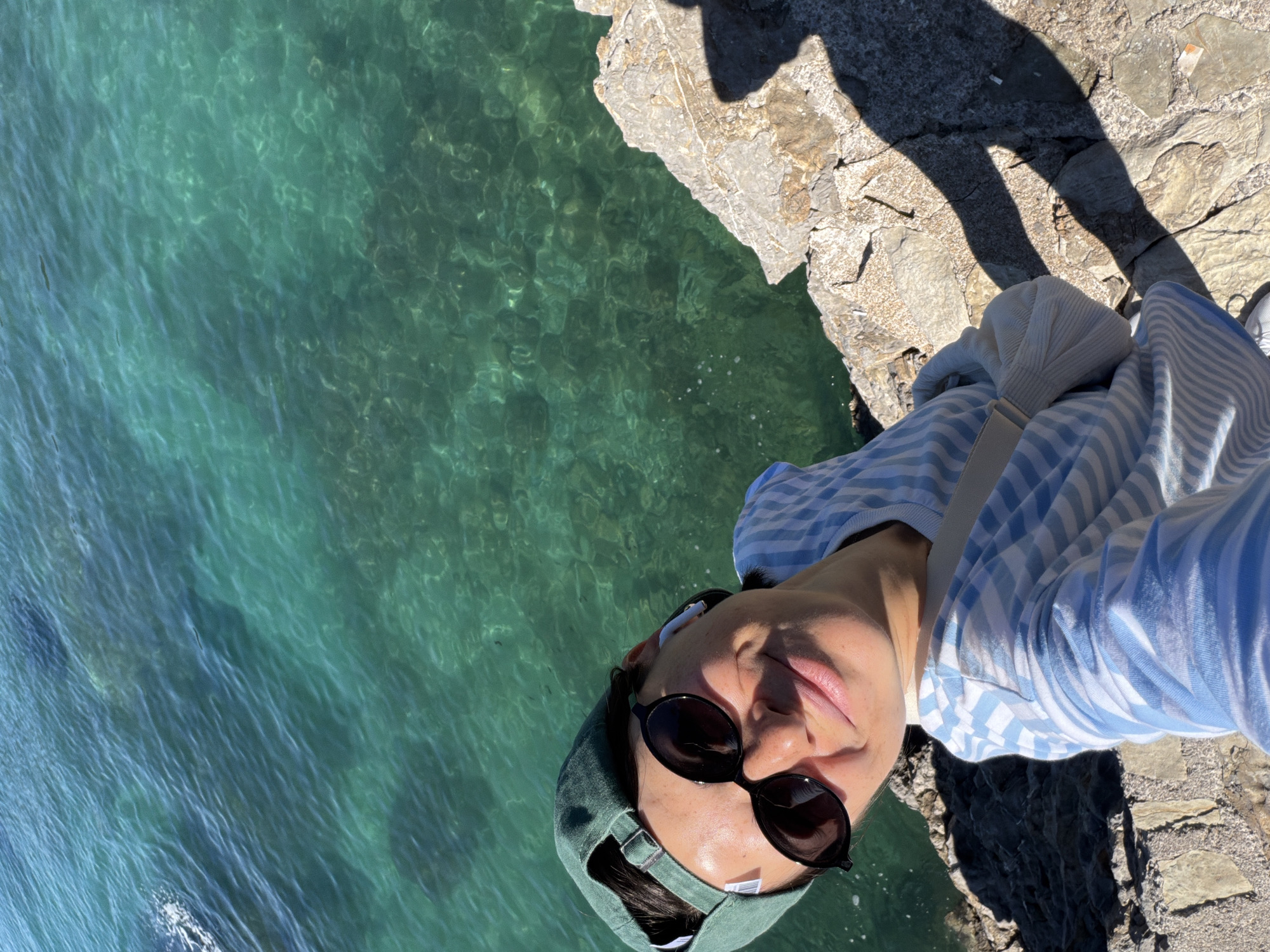 A person wearing sunglasses and a green baseball cap stands on a rocky outcropping next to clear, turquoise water. The person is dressed in a blue and white striped shirt. Part of a shadow of a person is cast on the rocky ground. The location appears to be a coastal area, possibly in Budva, Montenegro, based on the water color and architecture.