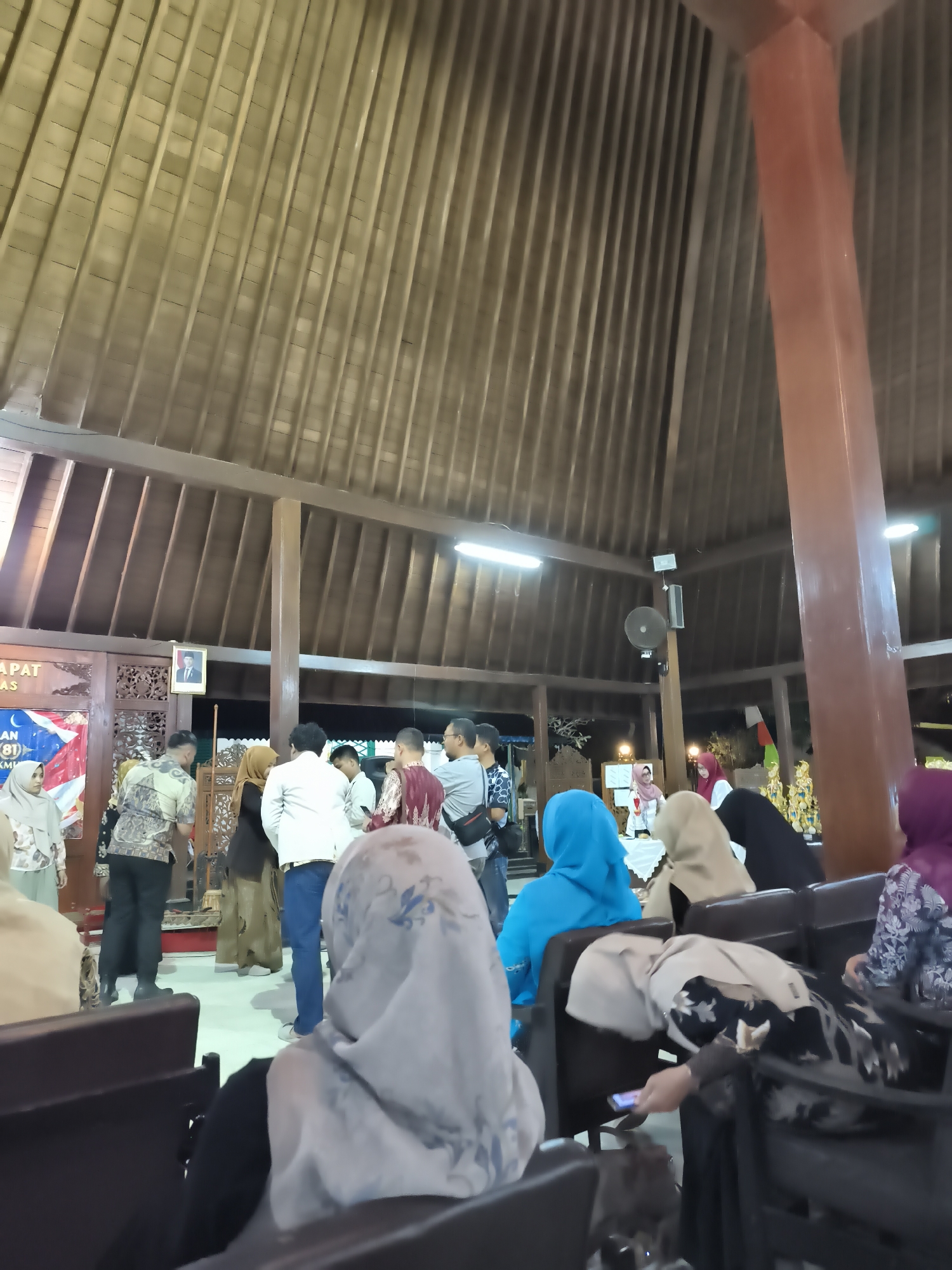 The image depicts an indoor gathering in what appears to be a hall with a high, sloped wooden ceiling supported by large wooden beams. Several people are present, mostly dressed in traditional attire, some wearing headscarves. In the background, there is a banner with text that is partially visible, and a framed portrait of a man in a suit hanging on the wall. A large, reddish-brown pillar stands prominently on the right side of the image. The setting suggests a cultural event or a community gathering in Banyumas, Indonesia. The foreground shows rows of dark seating, with some individuals already seated and others standing and interacting. One person in the foreground is looking at their phone.
