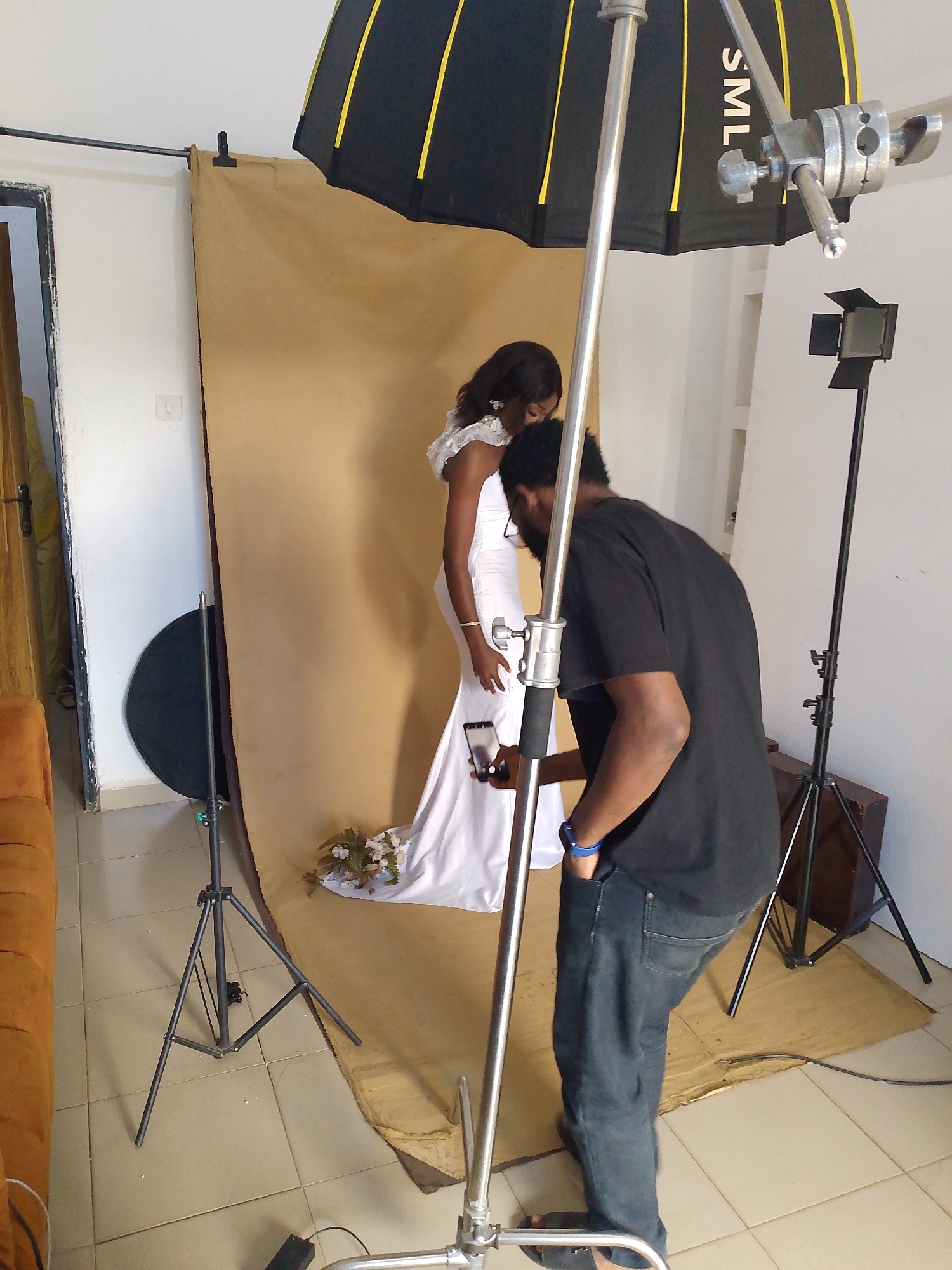 The image shows a photography studio setup with two people. A woman wearing a white dress, likely a wedding gown, stands facing away from the camera. She is posing against a tan backdrop. A man, possibly the photographer, is positioned behind her, looking at a phone he is holding. He is wearing a black t-shirt and blue jeans.
The studio is equipped with professional lighting equipment, including a large umbrella light positioned overhead and a smaller spotlight on a stand. There are also several light stands and a circular reflector visible in the scene. A section of an orange couch is visible on the left side of the image, and a wooden trunk is partially visible on the right. The floor appears to be tiled.
The overall impression is that of a photoshoot in progress, likely for a wedding or formal event. The lighting suggests an indoor setting, and there are no immediate cues about the time of day or weather. The individuals' focus on the task at hand indicates a professional or semi-professional environment. The text "SML" is visible on the umbrella light.