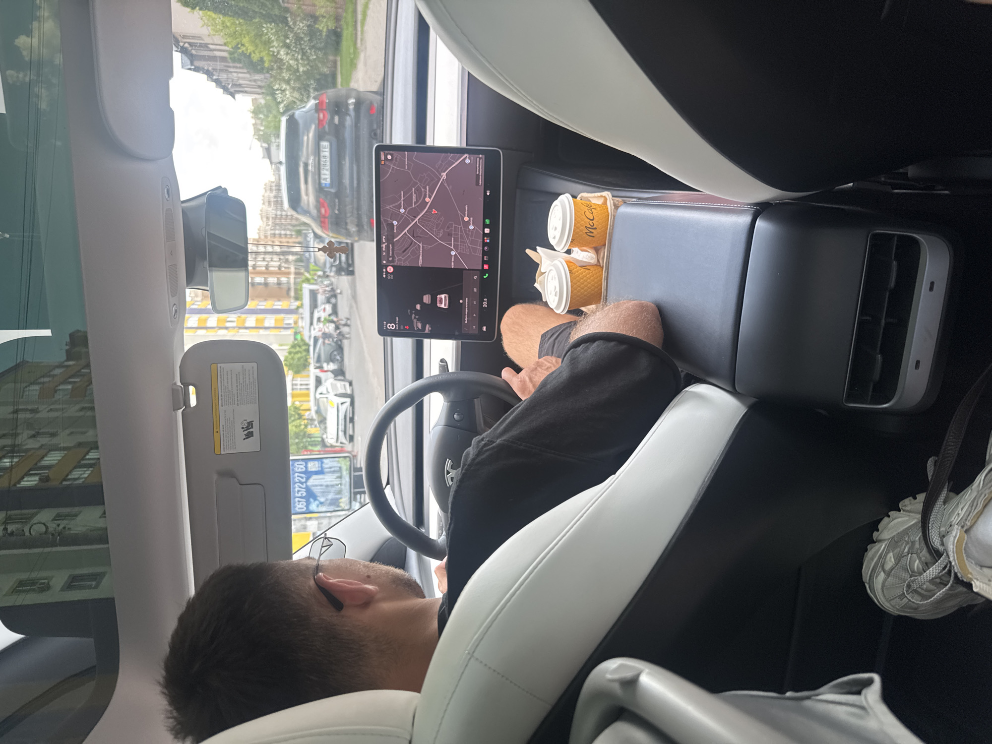 The image provides a view from the front passenger seat of a modern white and black Tesla car, looking towards the driver and the road ahead. A male driver, seen from behind, wears a dark shirt and glasses, with his forearms resting on his shorts-clad legs. He appears relaxed, possibly waiting in traffic or at a stop.
In the center console, two McCafé cups are placed in cup holders, suggesting the occupants are enjoying beverages. The car's prominent central touchscreen displays a map application showing an urban area, likely used for navigation. The screen also indicates "20.5" (presumably temperature in Celsius) and "8" (likely battery percentage). The steering wheel, partially visible, bears the Tesla logo (reflected on the screen). A small crucifix hangs from the rearview mirror.
Through the windshield, a city street is visible under a bright, daytime sky. Several cars are present, including a black SUV directly in front with the Ukrainian license plate "AI 2868 TE", which corresponds to Kyiv Oblast. Buildings with distinct architecture and trees line the street. A sun visor with visible warning labels is lowered slightly. On a distant billboard or bus stop, a Ukrainian phone number "067 572 27 60" is readable. In the bottom right, a foot wearing a light-colored sneaker is visible, belonging to a passenger. The setting is an urban environment, consistent with Sofiivska Borshchahivka, Ukraine.