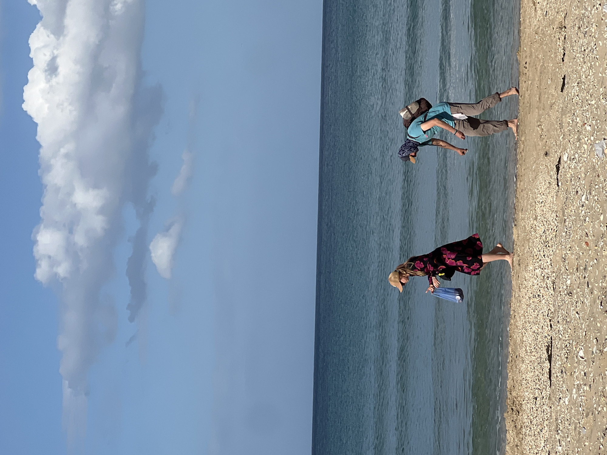 A vertical landscape depicts a sunny beach scene in M'Diq, Morocco. The left half of the image features a blue sky dotted with cumulus clouds. The right half shows a beach bordered by ocean. Two people are visible on the beach. A woman in a floral dress and straw hat walks along the shoreline carrying a blue plastic container. Further along the beach, a man wearing a backpack, khaki pants, and a blue shirt bends down near the water's edge. The beach is composed of sand and seashells. The water is calm.