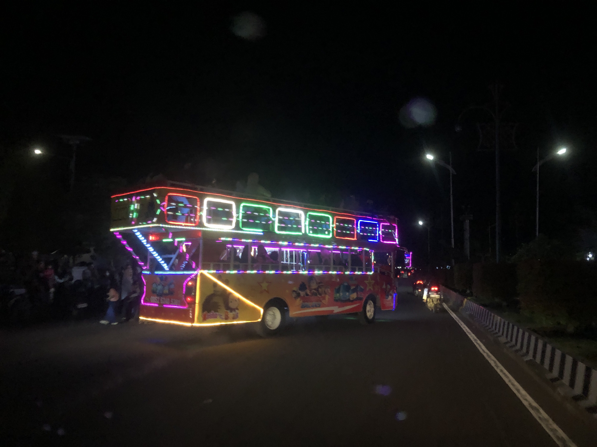 A brightly lit, open-top tour bus is parked on the side of a road in Lhokseumawe, Indonesia, at night. The bus is adorned with neon lights in red, green, white, blue, and pink, and it features cartoonish graphics on its lower panels. People are standing near the back of the bus. Streetlights illuminate the road, and there is a median with a white line. Other vehicles are visible further down the road.