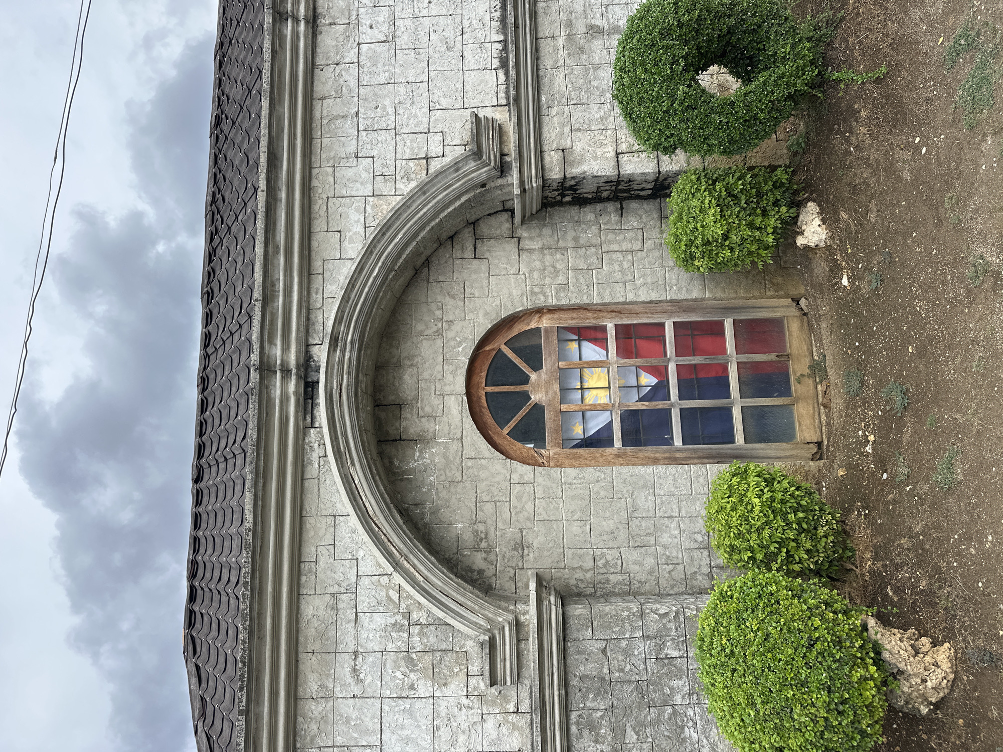 An outdoor, eye-level, medium shot captures a stone building facade in Oslob, Philippines, under an overcast sky. The building's exterior is constructed from light gray, rough-cut stones, arranged in a staggered pattern. A prominent arched architectural feature, also made of stone, is centered on the facade. Within this arch is a tall, narrow window with a wooden frame and divided panes. The window glass is tinted with the colors of the Philippine flag: red, blue, and yellow, creating a patriotic stained-glass effect. The roof above the facade is covered in dark, wavy tiles. In front of the building, on the ground, are three manicured, spherical green bushes. Two are smaller and positioned to the left of the window, while a larger one is to the right. The ground is covered in dirt, small stones, and sparse dry vegetation. Two power lines are visible in the upper portion of the image, stretching across the cloudy sky.