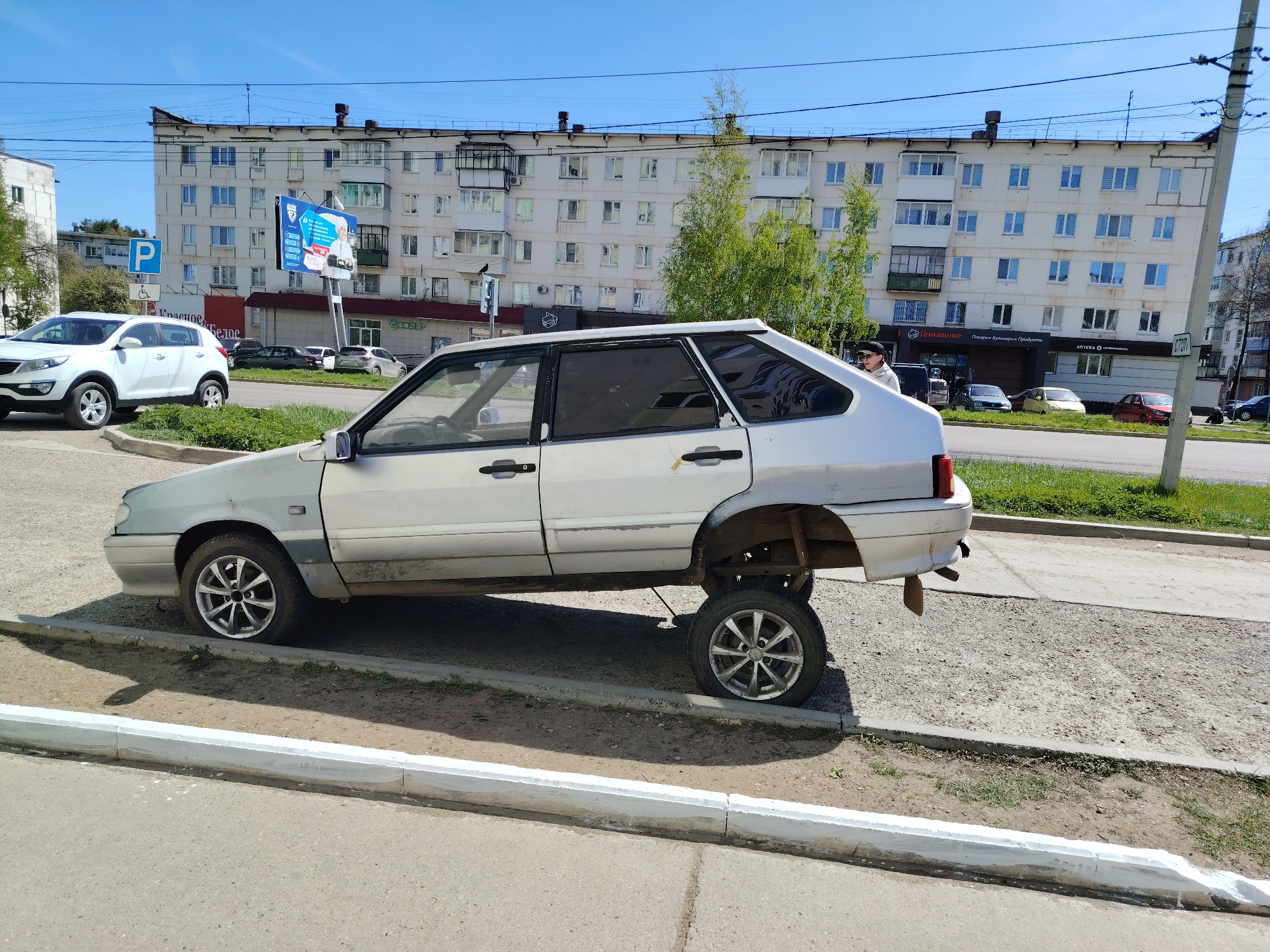 A silver Lada Samara automobile, missing its rear passenger tire, is awkwardly perched atop the curb of a sidewalk in Chaikovsky, Russia. The missing tire has been placed under the automobile near the rear, to possibly support the car. The sidewalk is constructed of concrete and is partially surrounded by a low white-painted border. In the background is a multi-story residential apartment building with a blue and white advertising billboard. Several other cars and pedestrians can be seen in the vicinity. The sky is clear and blue, indicating daytime.