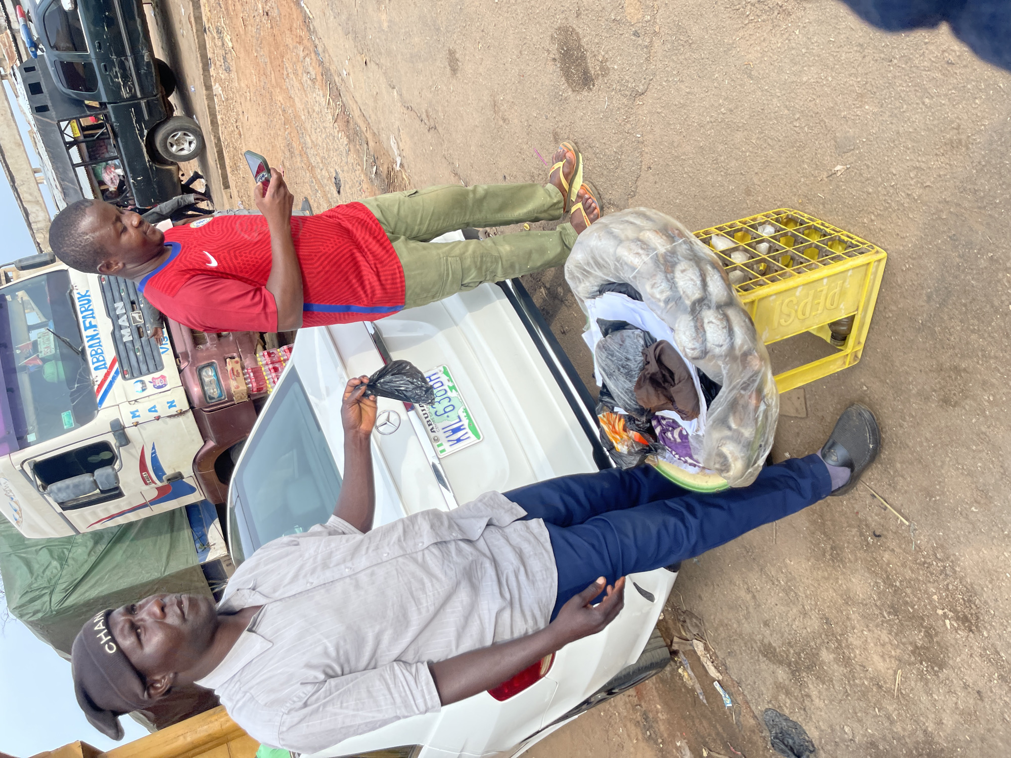 In Jos, Nigeria, two men stand beside a white sedan. The man on the left, wearing a grey shirt and dark beanie, holds a black plastic bag. In front of him, a collection of clothing is displayed in a clear plastic bag, resting on a yellow Pepsi crate. The man on the right, in a red shirt and green pants, looks at his phone. The background features a large truck and a smaller pickup truck, suggesting a roadside or market setting. The ground is dusty earth, indicative of an outdoor environment.