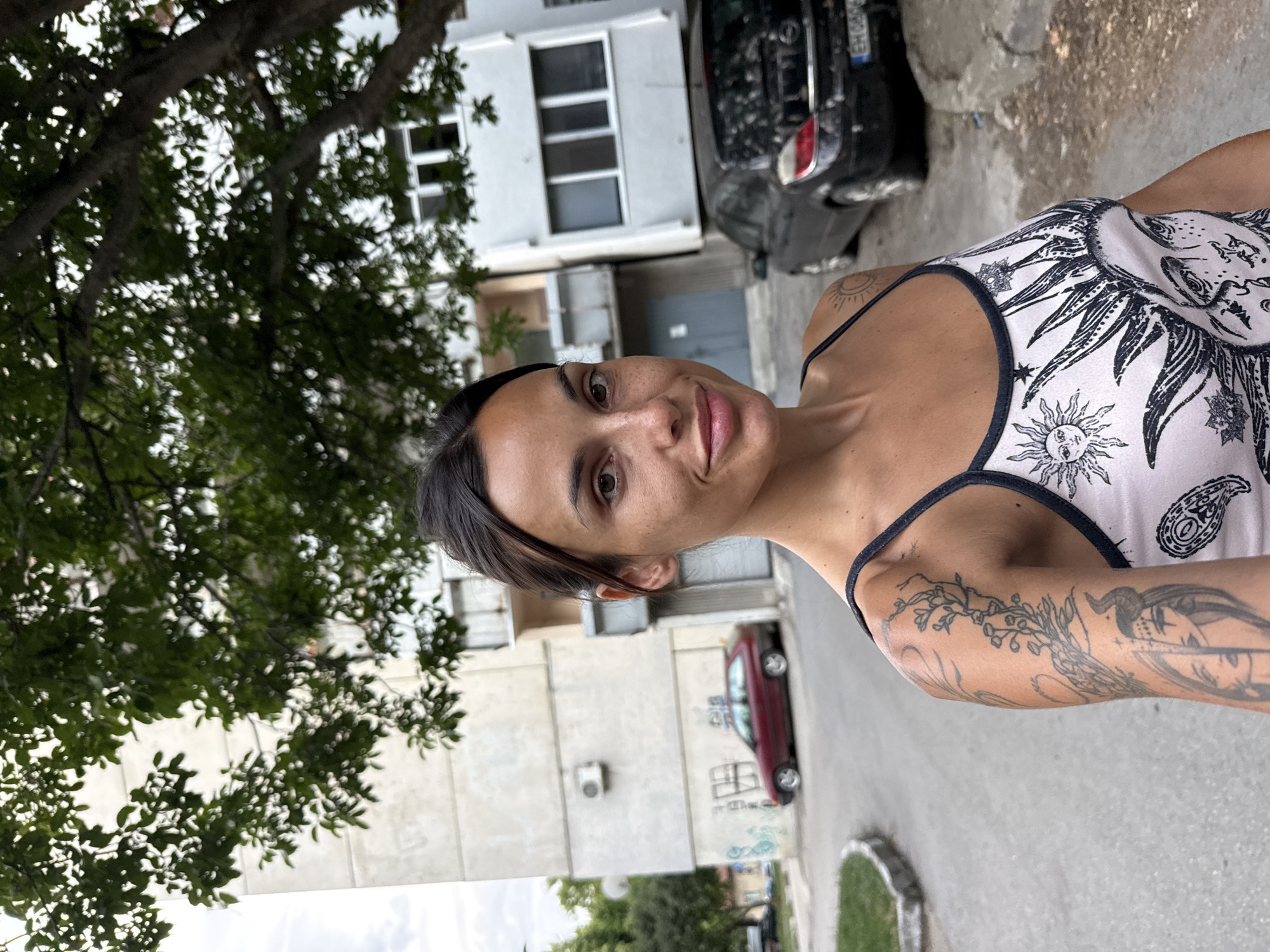 A woman with dark hair, freckles, and tattoos on her arm is standing outside an apartment building in Pleven, Bulgaria. She is wearing a white tank top with a sun design. Behind her are several apartment buildings, a red car, and a black car. A tree with green leaves is in the foreground.