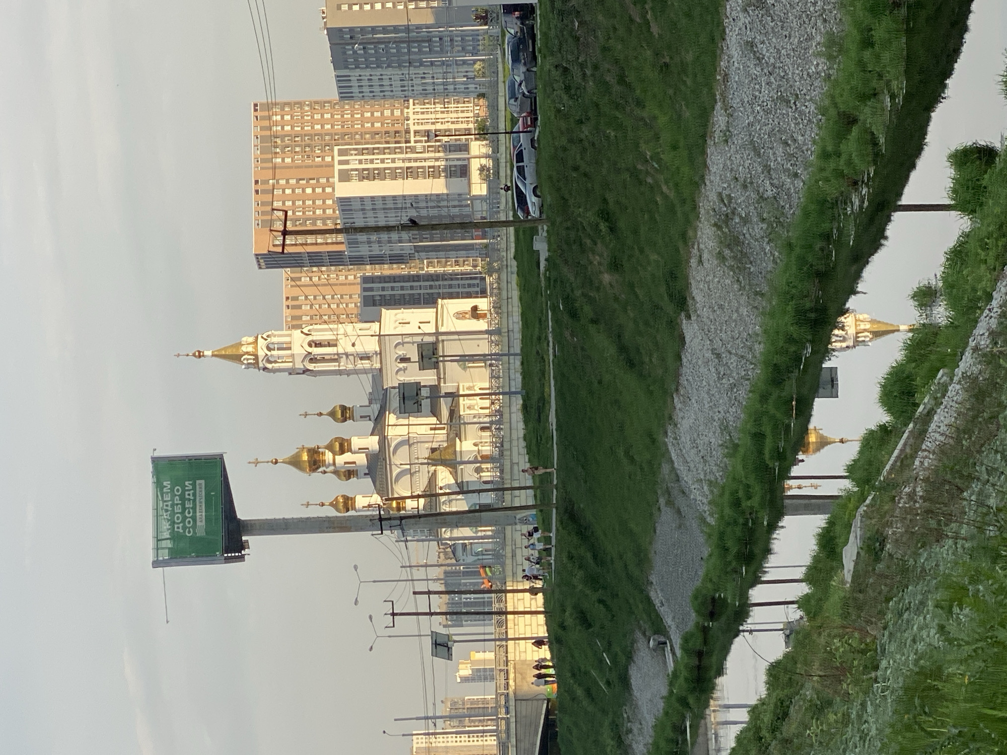 The image captures a scene in Yekaterinburg, Russia. A Russian Orthodox church with golden domes dominates the left side, juxtaposed against modern high-rise buildings in the background. A billboard with Russian text stands near the church. A group of people are visible along a path beside what appears to be a water body, which reflects the buildings and church, creating a mirrored effect. The scene is captured in daylight, likely late afternoon. A grassy area with gravel borders the water's edge. Power lines and lampposts line the street near the church.