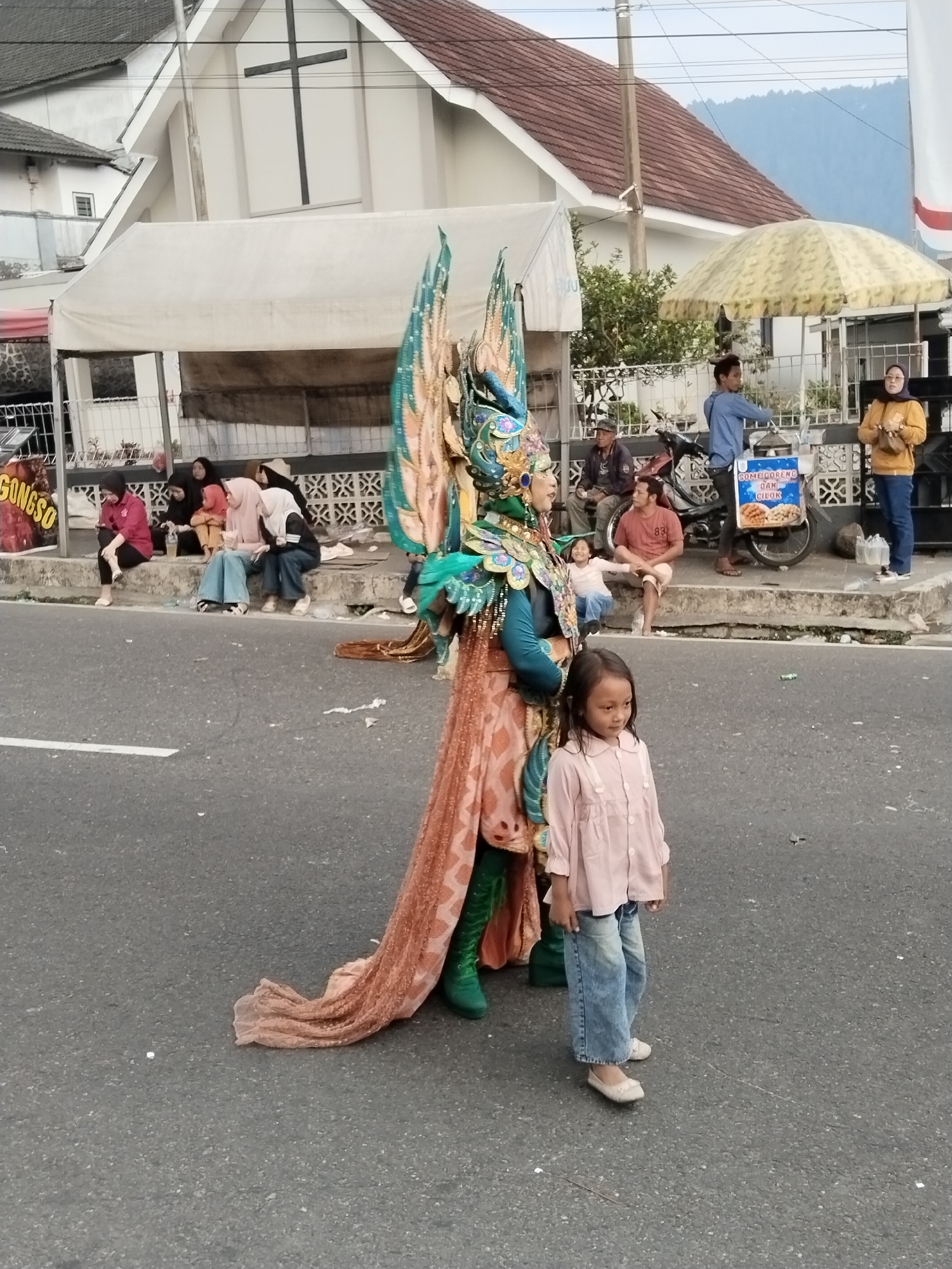 A person in an elaborate costume, featuring large feathered wings and intricate headdress, stands on a street in Nglebak, Indonesia. The costume is primarily teal and gold, with a flowing orange and pink fabric skirt. To the right of the costumed person is a young girl wearing a pink shirt and blue jeans, looking to her left. In the background, several people are seated on a curb, some watching a parade or event. A white tent is set up behind them, and further back, a building with a red-tiled roof and a black cross is visible. A street vendor's cart is also present, with an umbrella offering shade. The street is paved and has a white dividing line.