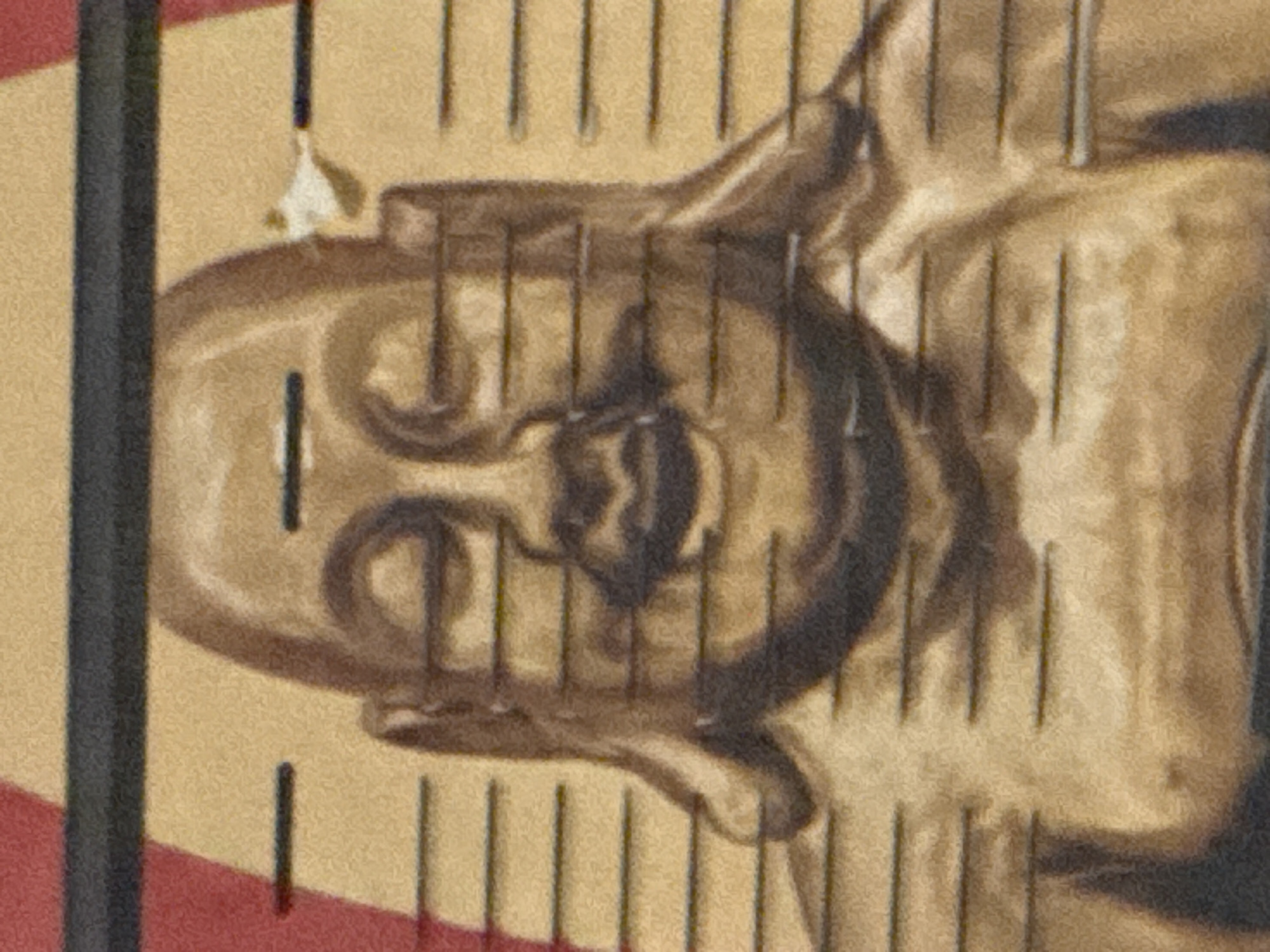 The image is a close-up, horizontally oriented, grainy photograph of a golden Buddha statue. The statue is depicted from the chest up, with its face prominently featured. Horizontal lines, resembling a measuring scale or a pattern, are superimposed across the image, passing over the statue. The background appears to be a textured yellow, with a sliver of red visible at the top left corner. There is a dark, thick horizontal band across the very top of the image. A small, irregular white object, possibly a piece of paper or a tag, is attached to the top right side of the statue's head. The statue's features, including its closed eyes, rounded cheeks, and serene expression, are visible. Its hands are not fully visible in the frame. The overall lighting is subdued, giving the statue a warm, aged appearance. There is no indication of the city of Shkotovo, Russia, within the image itself.