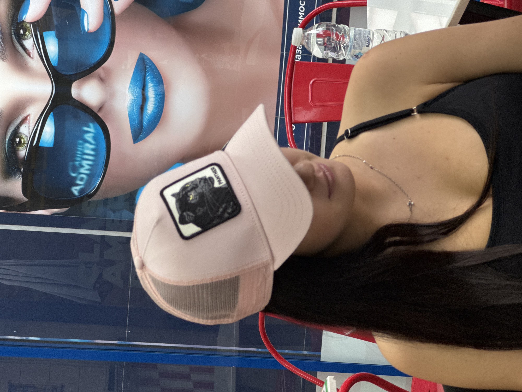 A young woman with dark hair, wearing a black tank top and a pink trucker hat featuring a panther patch, is seated indoors. She has a silver necklace and is positioned near a red trolley. In the background is an advertisement featuring a woman wearing blue sunglasses and lipstick with the words "ADMIRAL" in the lens. The background also suggests a location in Asenovgrad, Bulgaria, based on visual cues. A bottle of water and white napkins are on the trolley.