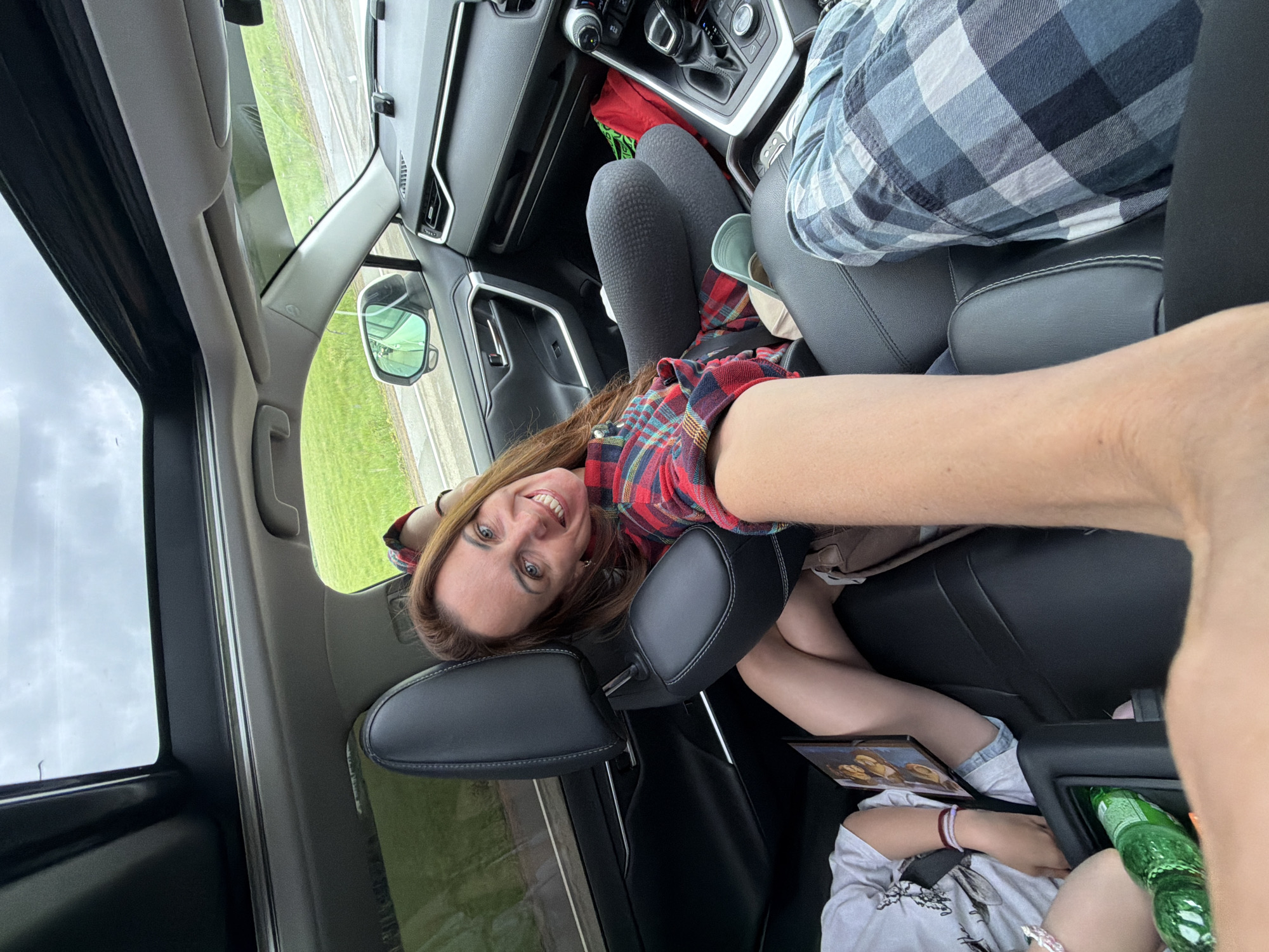 In a car's interior, a woman with long brown hair and blue eyes smiles at the camera while holding it in a selfie position. She is wearing a red and black plaid shirt and dark gray leggings. Her legs are crossed, and she is leaning back against a black leather car seat. To her left, a child wearing a white t-shirt and denim shorts sits with their legs crossed and a tablet resting on their lap. A green bottle of soda is visible in the foreground. To the right of the woman, another person is partially visible, wearing blue and gray plaid pants. The car has a panoramic sunroof, and through the windows, a grassy field and cloudy sky are visible. The setting appears to be a road trip, possibly on the outskirts of a city like Kirga, Russia, given the natural landscape outside.