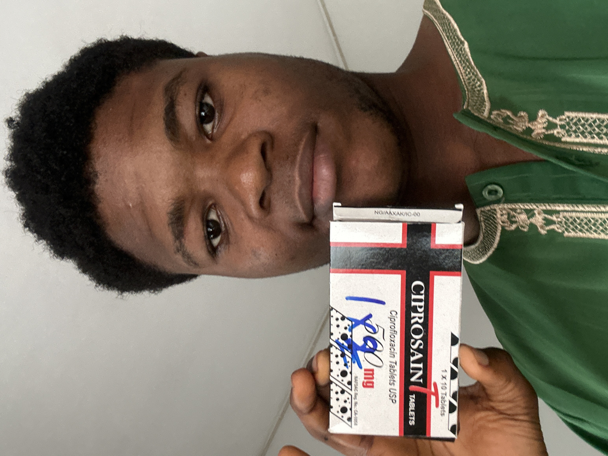 A young Black male is holding up a small, rectangular box of medication, possibly Ciprofloxacin tablets, to the camera. The box is white and red, with black text. On the box, there is a handwritten blue inscription that appears to be a dosage or quantity. The man is wearing a green garment with gold embroidery around the neckline. He has short, dark, curly hair and is looking directly at the camera with a neutral expression. The background is a plain, light-colored wall, suggesting an indoor setting. The image quality is good, with clear focus on the man and the medication box. The lighting is even, indicating no strong light sources. The image does not provide any information that can identify the location as Ihiagwa, Nigeria.