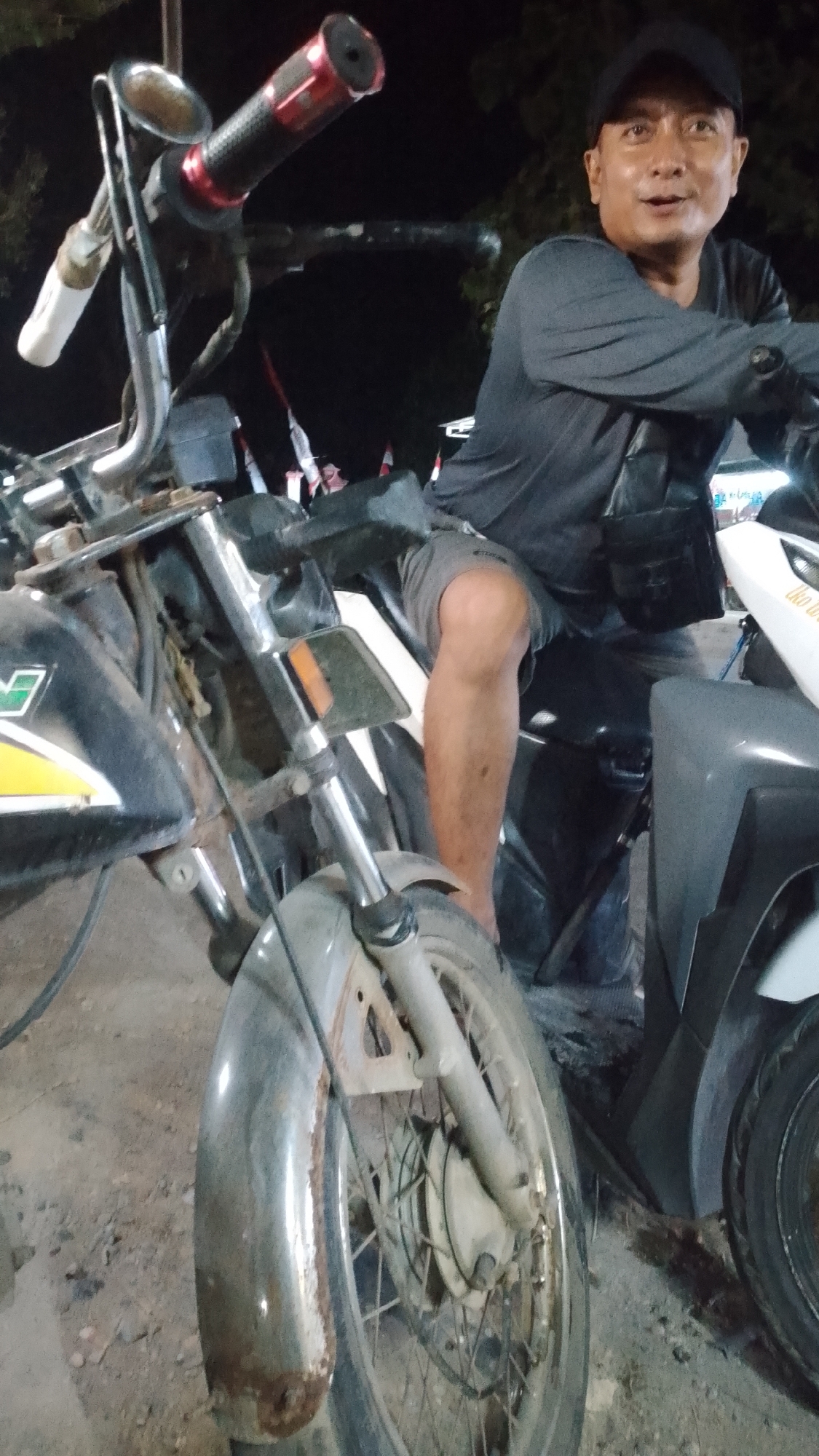 A man wearing a black baseball cap and a dark long-sleeved shirt and grey shorts is sitting on a motorcycle in what appears to be a street at night. He is looking towards the camera and smiling slightly. The motorcycle is black and has a chrome front fender. The man has a dark bag slung over his shoulder. In the background, there are red and white flags, possibly national flags, and some trees. The ground appears to be dirt or gravel. The lighting suggests artificial illumination, likely from streetlights or nearby buildings. The image was taken in Karanglangu, Indonesia.