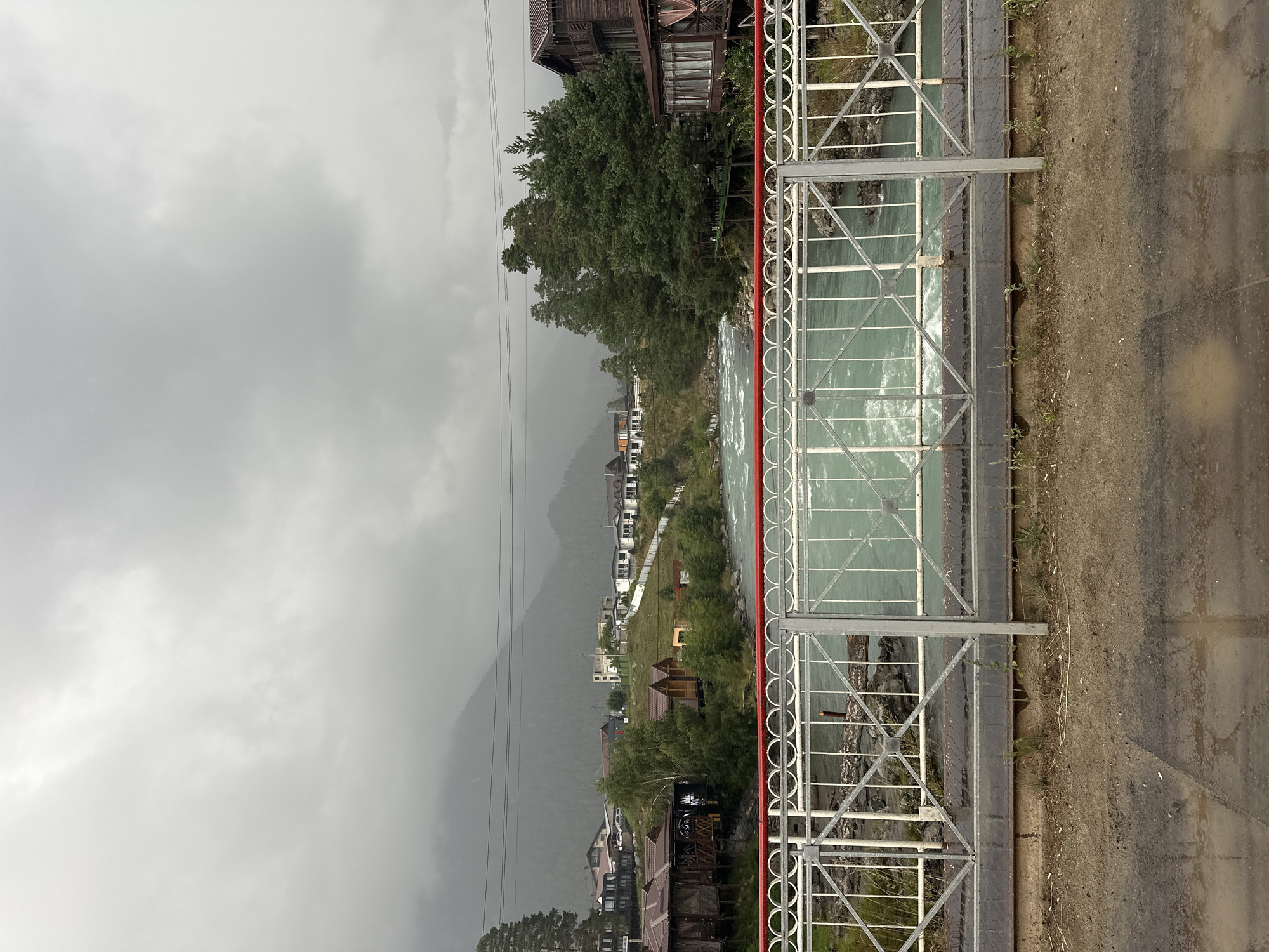 This is a medium-shot, eye-level, outdoor, still photograph taken on a cloudy day. The viewpoint is from a bridge, looking across a river towards a settlement nestled at the base of a mountain. In the foreground, a metal railing with a white lattice pattern and a red top rail is visible. The railing is supported by grey vertical posts. Beyond the railing, a wide, turbulent, turquoise river flows rapidly. The river is bordered by rocky banks and lush green vegetation. On the far bank of the river, a cluster of wooden buildings with dark roofs, some appearing to be cafes or restaurants with outdoor seating, are situated. Further up the slope, a row of white buildings with darker roofs forms a residential area. Behind the settlement, a dense, dark green pine forest covers the lower slopes of a large, mist-shrouded mountain. The sky is overcast with grey clouds, obscuring the upper reaches of the mountain. The overall scene depicts a scenic, mountainous landscape with a river flowing through a small settlement in Arkhyz, Russia. There are no people visible in the image.