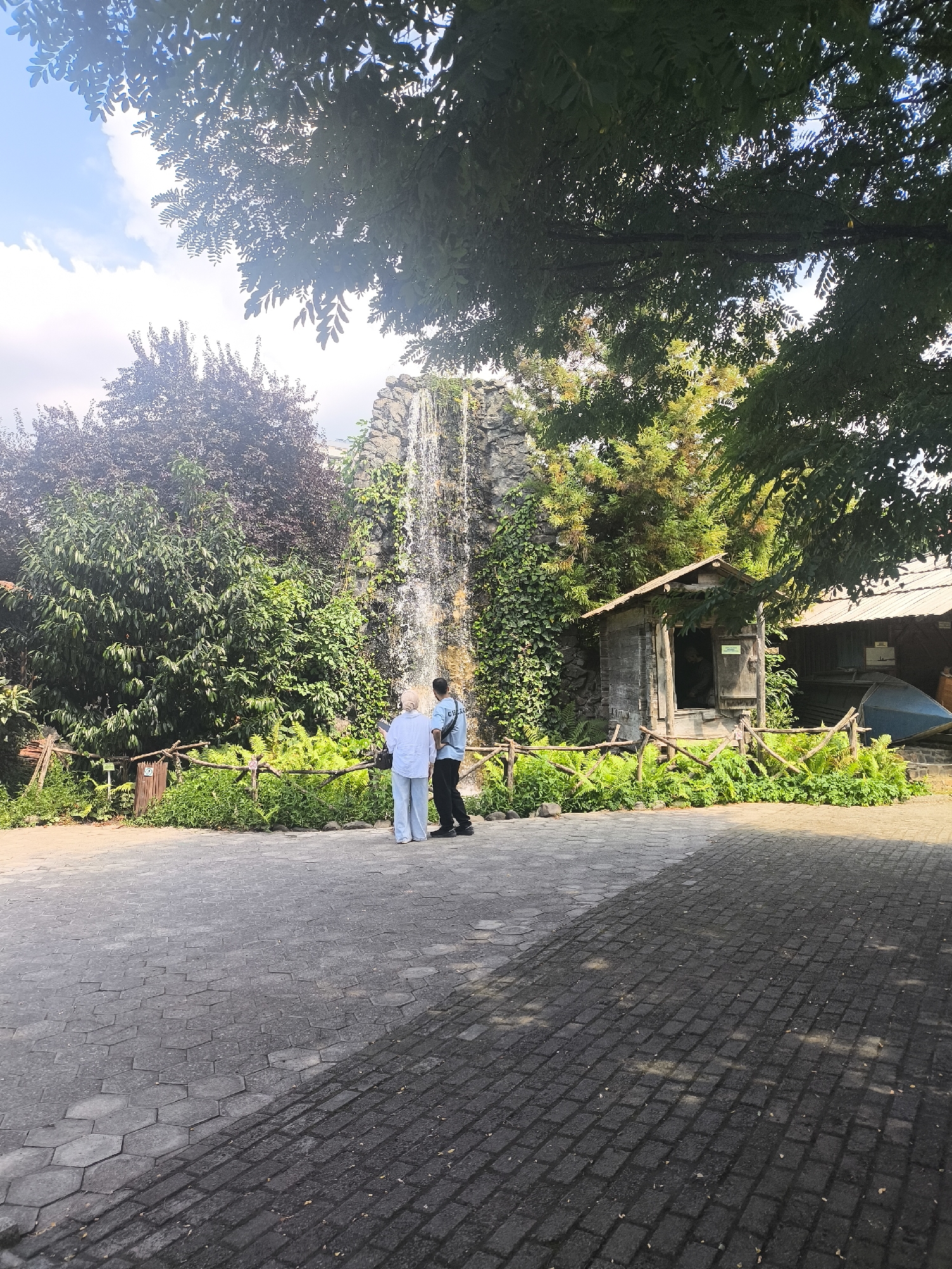 The image shows an outdoor scene with a waterfall cascading down a rocky surface covered in greenery. In front of the waterfall, two individuals, one wearing a light blue shirt and light-colored pants, and the other in a dark shirt and black pants, are standing and looking towards the waterfall. A small, rustic wooden structure with an open doorway is visible to the right of the waterfall, partially obscured by trees. A wooden fence with rough-hewn posts and branches encloses an area with lush green plants in front of the waterfall and the wooden structure. The ground in the foreground is paved with interlocking hexagonal and rectangular gray stones, with shadows indicating a sunny day. Trees with dark green and reddish-brown foliage are present on the left and overhead, with some branches extending into the frame. The sky is visible in the upper part of the image, with scattered white clouds. There are no specific elements in the image that definitively place it in Batumi, Georgia.
