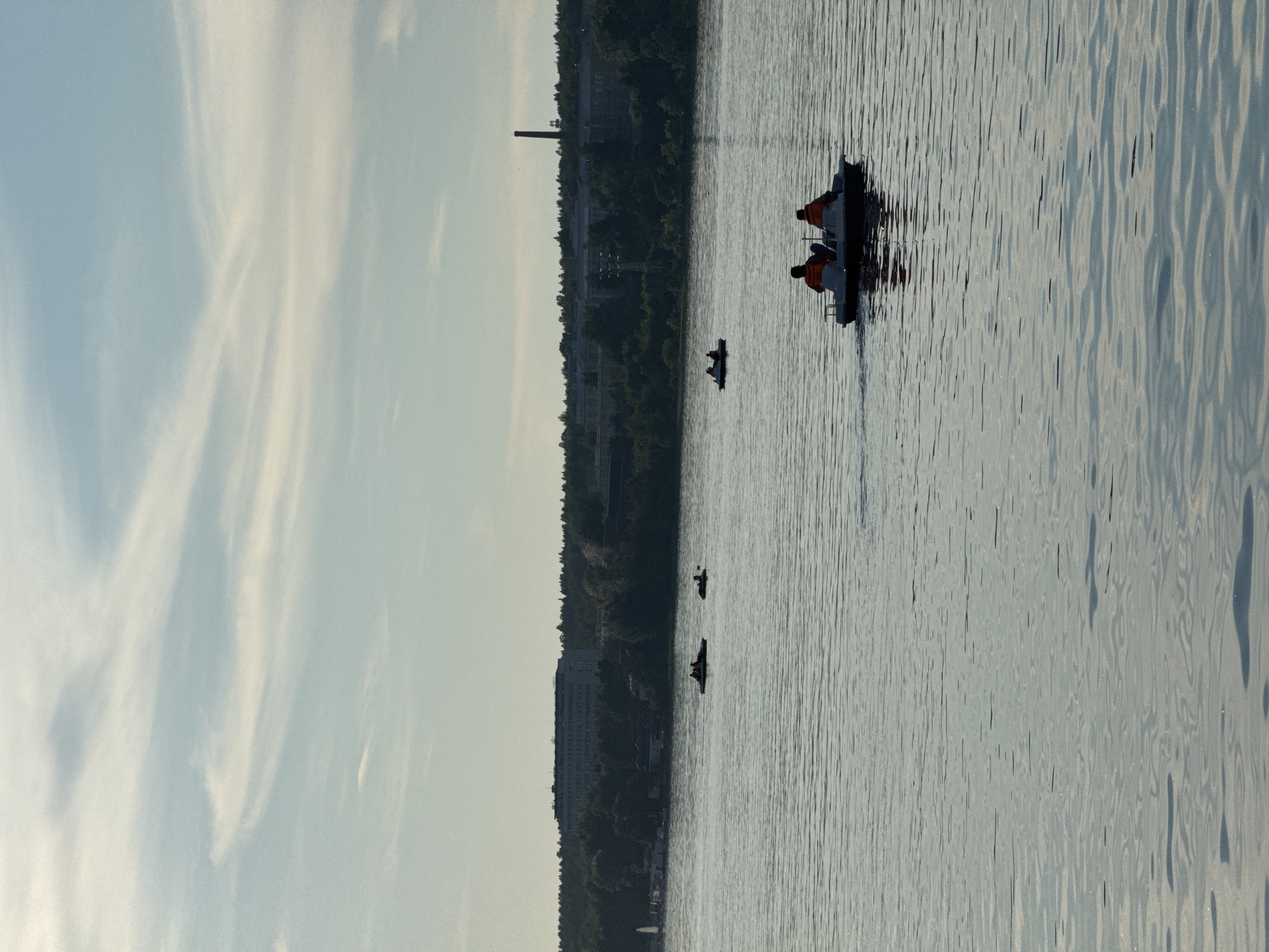 A wide shot captures a calm body of water, likely a reservoir or large river, under a pale blue sky with wispy white clouds. In the foreground, the water is rippled, reflecting the muted light. Several small boats are scattered across the water. In the middle distance, a pedal boat with two individuals wearing orange life vests is moving away from the viewer. Further back, two other small boats are visible, each with at least one person. The background features a densely wooded shoreline, beyond which lies a long, multi-story building that appears to be a complex or factory, with a tall, slender smokestack visible to its right. The overall scene suggests a leisurely day on the water, with distant urban or industrial elements. This setting is consistent with the shoreline of the Izhevsk Reservoir in Izhevsk, Russia, a popular recreational area.