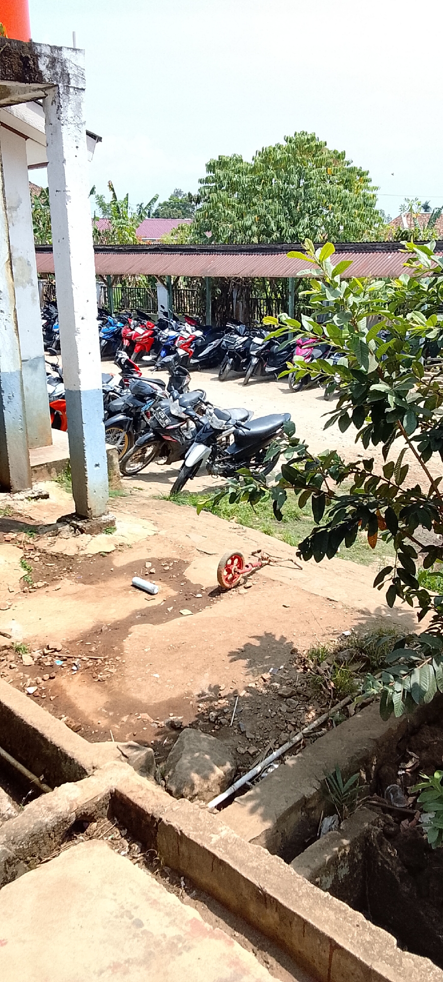 The image shows a parking lot for motorcycles in what appears to be a school or public facility in Jepara, Indonesia. The lot is filled with numerous motorcycles parked closely together under a covered structure with a corrugated metal roof. The structure is supported by concrete pillars, one of which is in the foreground and shows signs of weathering. The ground of the parking lot is a mixture of dirt and concrete. In the foreground, there is a broken red toy with a single wheel and a white cylindrical object lying on the ground. Beyond the parking lot, there are trees and some buildings with red roofs, typical of Indonesian architecture. The sky is bright and overcast, suggesting daytime. There are no people visible in the image. The scene depicts an organized but somewhat cluttered outdoor space, likely a daily scene at a busy location in Jepara.