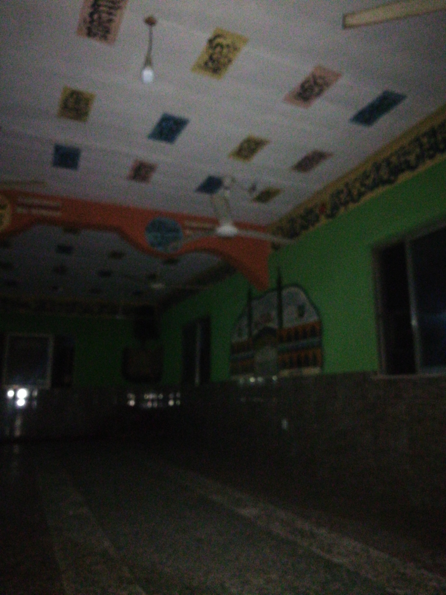 This dimly lit, blurred image depicts the interior of what appears to be an Islamic prayer hall or mosque. The ceiling is light-colored, adorned with multiple square and rectangular panels in varying hues like yellow, blue, black, and red/orange, some of which feature faint, illegible Arabic calligraphy. A single, illuminated light bulb hangs from the ceiling in the upper-center, and a blurry ceiling fan is visible further back.
The upper sections of the walls are painted a vibrant green, contrasting with a darker, possibly tiled or paneled, lower section. Along the top of the green wall, just below the ceiling, runs a decorative band containing intricate patterns or script that is also indistinct. An arched, orange and red decorative border with a blue circular emblem extends across the upper left wall. Further right, a larger, stylized silhouette of a building, reminiscent of a mosque with domes and minarets, is visible against the green wall. Two dark, rectangular areas on the right wall suggest windows, through which no external light or view is discernible, indicating it might be evening or night.
The floor is dark and appears to be covered with patterned carpet or tiles. No people or specific activities are present, leaving the room empty and conveying a sense of quietude or contemplation. The overall low light levels suggest the photograph was taken in a dimly lit environment, possibly at night.