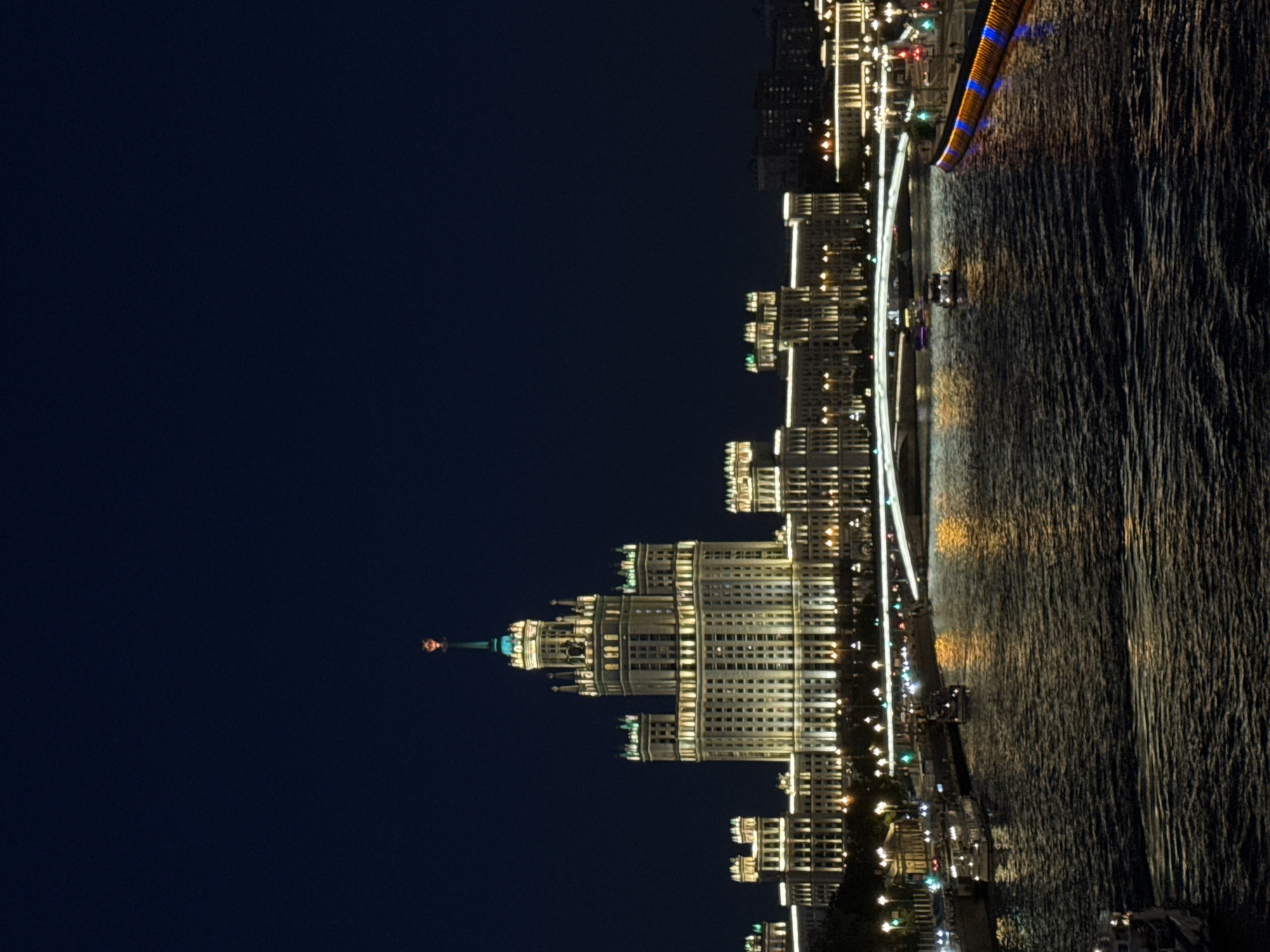 This image captures a nighttime cityscape along the Moskva River in Moscow, Russia. The central and most prominent feature is the brightly illuminated Kotelnicheskaya Embankment Building, a Stalinist skyscraper with multi-tiered architecture, topped by a green spire bearing a faint red star at its peak. The building's facades are uniformly lit with warm white and yellow lights, highlighting its details.
The Moskva River occupies the lower half and midground of the frame, its dark, textured surface reflecting the numerous city lights in elongated, shimmering patterns. Several small boats or ferries are visible on the river; some are docked along the left embankment, while others appear to be moving in the central waterway, including one passing under a white, arched bridge that spans the river in the middle distance. This bridge is also illuminated with white light.
The riverbanks are lined with other buildings, some featuring similar architectural styles and illumination as the main skyscraper, as well as a series of streetlights that emit white and green light. On the right embankment, a section of the river wall displays a distinct blue and orange linear illumination. The sky above is a deep, clear navy blue to black, providing a stark backdrop to the brightly lit urban scene. No discernible individuals or specific human interactions are visible within the frame.