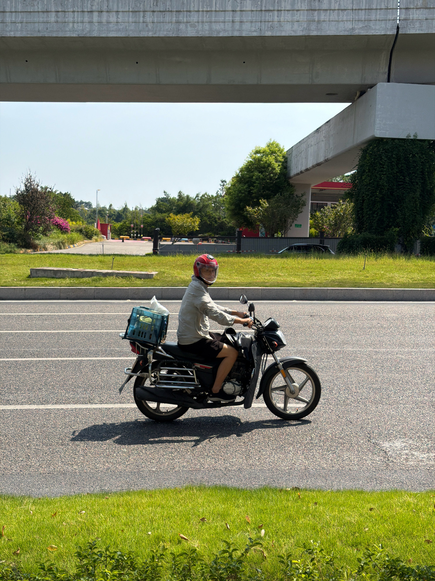 A person wearing a red helmet and a light-colored shirt rides a motorcycle on a paved road in Jiangjin, China. The motorcycle has a blue plastic crate attached to the rear, containing a white plastic bag. The road is bordered by a grassy area and an elevated concrete structure overhead. In the background, there are trees, some buildings, and a parking lot. The sky is clear and blue.