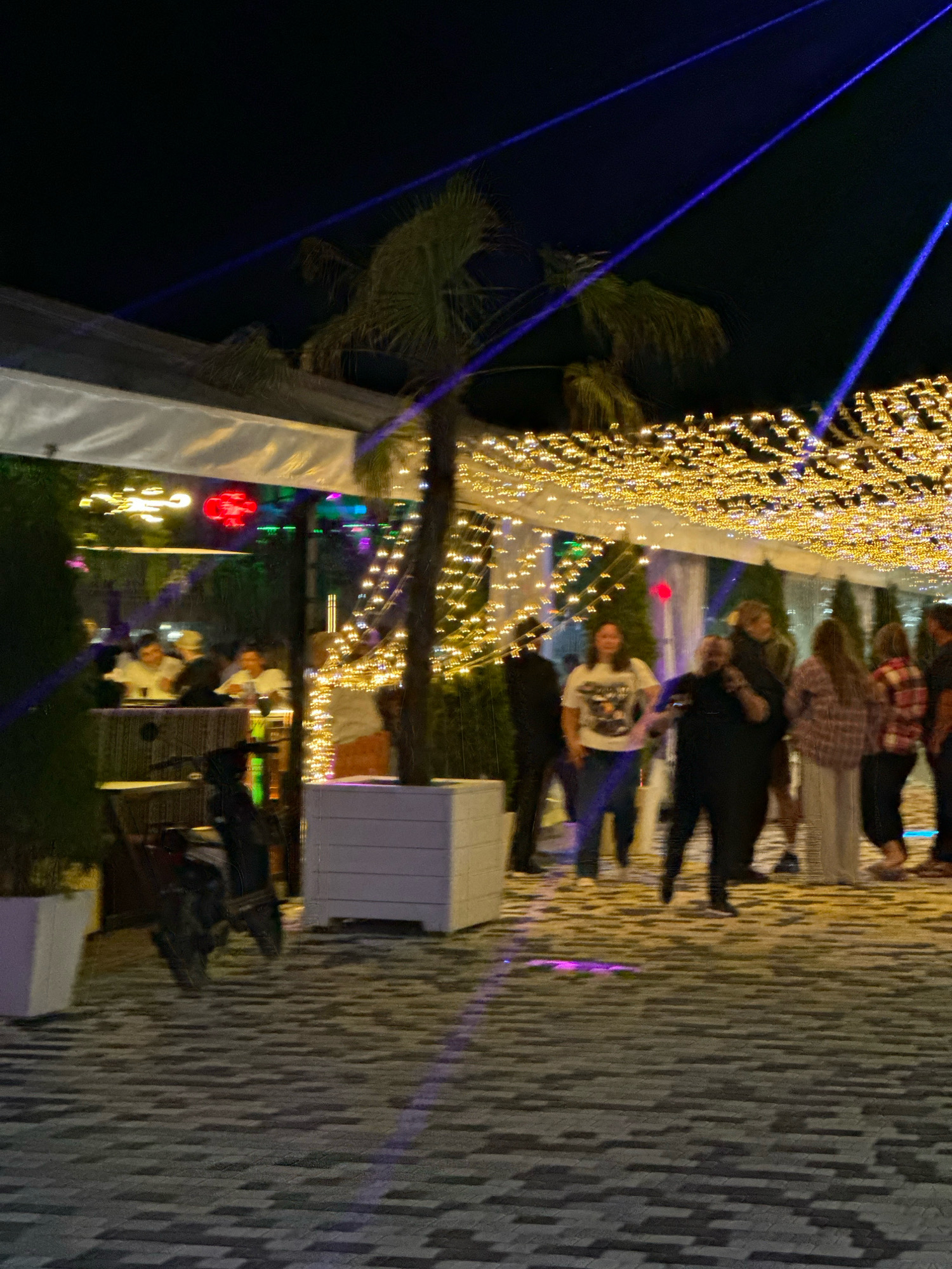The image depicts an outdoor scene at night, possibly a restaurant or event venue, in Lermontovo, Russia. Blue laser beams are projected across the dark sky, intersecting with strings of warm-toned lights draped overhead. A palm tree stands near the center, illuminated by the festive lighting.
In the foreground, a paved walkway made of dark and light grey interlocking bricks leads into the venue. To the left, a dark motorcycle or scooter is parked, partially obscured by a green bush and a white planter.
Several people are visible in the mid-ground and background. Some individuals are seated at tables within the covered area, which is brightly lit with colorful lights, including a prominent red circular light. Others are standing and walking on the paved area. One person in the foreground appears to be walking towards the viewer, while others are seen socializing or moving away. The people are dressed in casual attire. The overall atmosphere suggests a lively evening gathering.