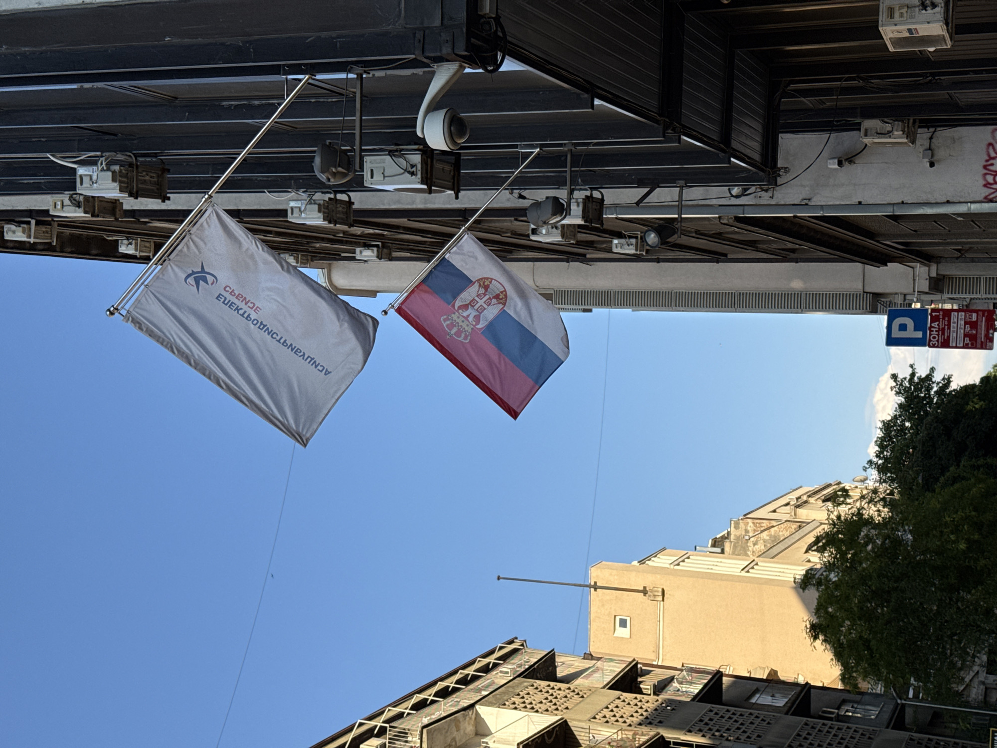 The image captures a street scene in Belgrade, Serbia, likely taken from a low angle looking upwards. Two flags are prominently displayed: the flag of Serbia (red, blue, and white horizontal stripes with the coat of arms) and another flag with text in Cyrillic, likely the flag of Crna Gora. The flags are hanging from metal poles attached to a building facade. The building is equipped with security cameras and lighting fixtures. Street signage is visible, including a parking sign. In the background, there are buildings with various architectural styles and trees. The sky is clear and blue.
