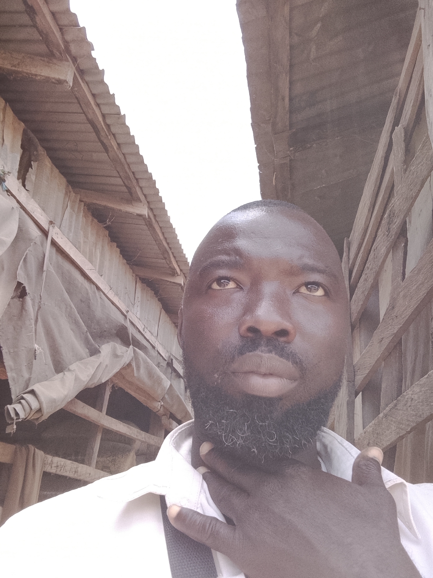 A man with dark skin and a full beard is positioned in a narrow alleyway with corrugated metal structures and wooden slats overhead. He is wearing a white shirt and has one hand on his throat. The environment is weathered and appears to be in an urban area. No specifics about the city of Jos, Nigeria can be ascertained from the image.