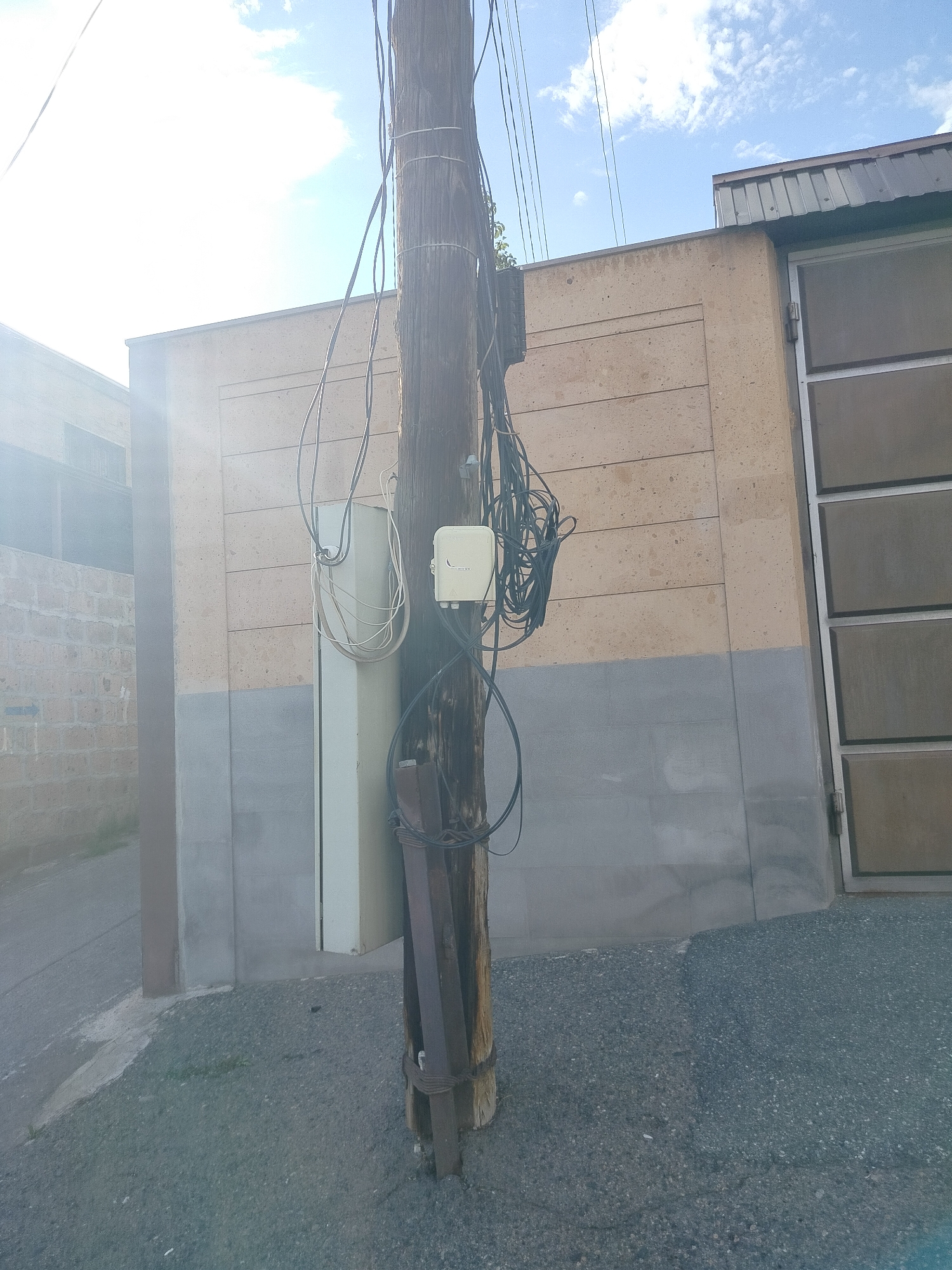 A wooden utility pole stands in the foreground, surrounded by various wires and junction boxes. A white cabinet is attached to the pole. The base of the pole is reinforced with metal braces. Behind the pole is a building with a two-tone beige and gray exterior. A gray door is on the right side of the building. The ground is paved. The image appears to be taken in Yerevan, Armenia, due to the architectural style and the type of utility infrastructure present. The sky is visible in the background, with some clouds present. The sun is shining, creating a bright and somewhat overexposed appearance.