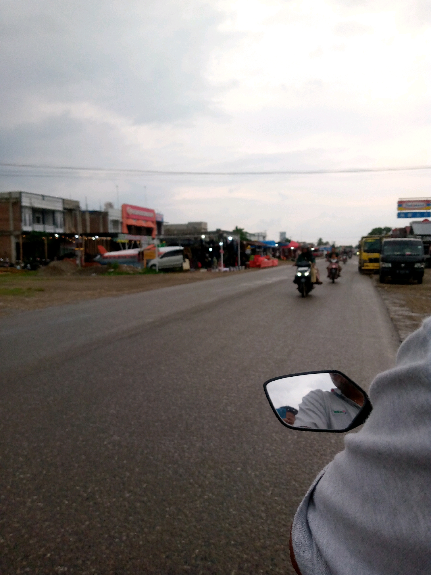 The image depicts a street scene in Gampongpaya, Indonesia, under an overcast sky. In the foreground, a person's arm and shoulder, clad in a grey garment, are visible. Attached to the side of a vehicle, likely a motorcycle, is a side-view mirror reflecting the driver and a portion of the sky. The road is wet, suggesting recent rain. On the left side of the road, buildings with storefronts are lined up. One building features an unusual display: a miniature airplane and a white car parked nearby. Further down the road, several motorcycles are in motion, with riders visible on some. On the right side of the road, a yellow truck and a black truck are parked. The scene appears to be a commercial or market area with ongoing activity.