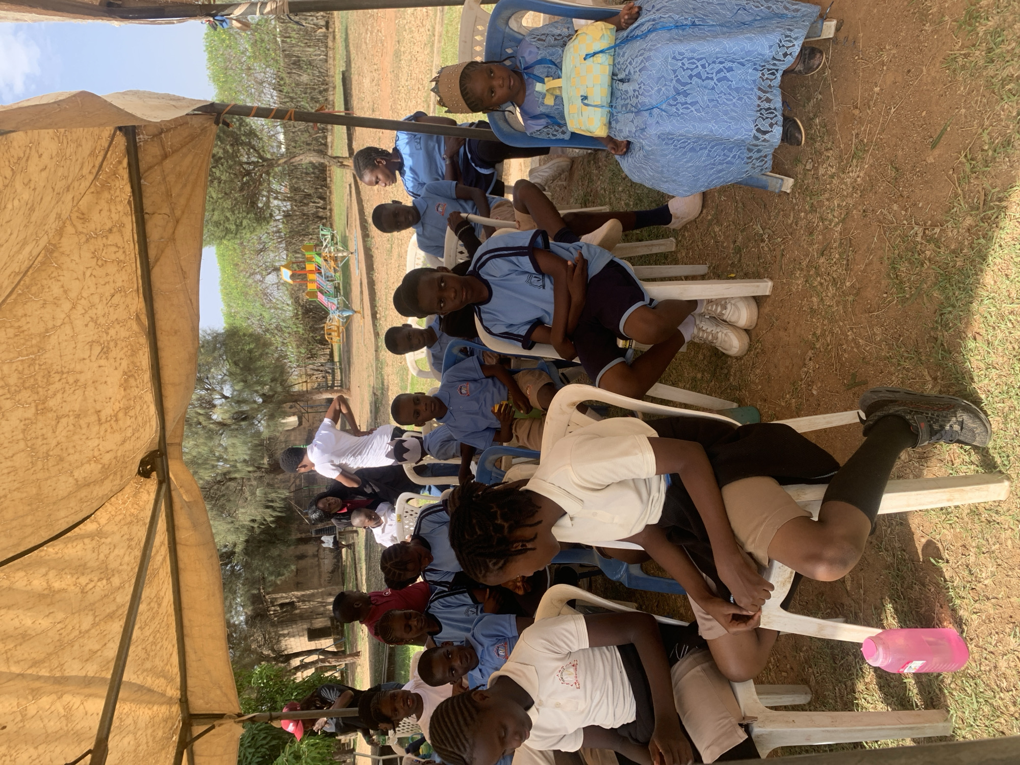 A group of children and adults sit on white plastic chairs under a tan tent in Jos, Nigeria. The children are wearing school uniforms and the adults appear to be teachers. The tent shields them from the sun, which is high in the sky. In the background there are trees and a colorful playground. The ground is dirt and grass.