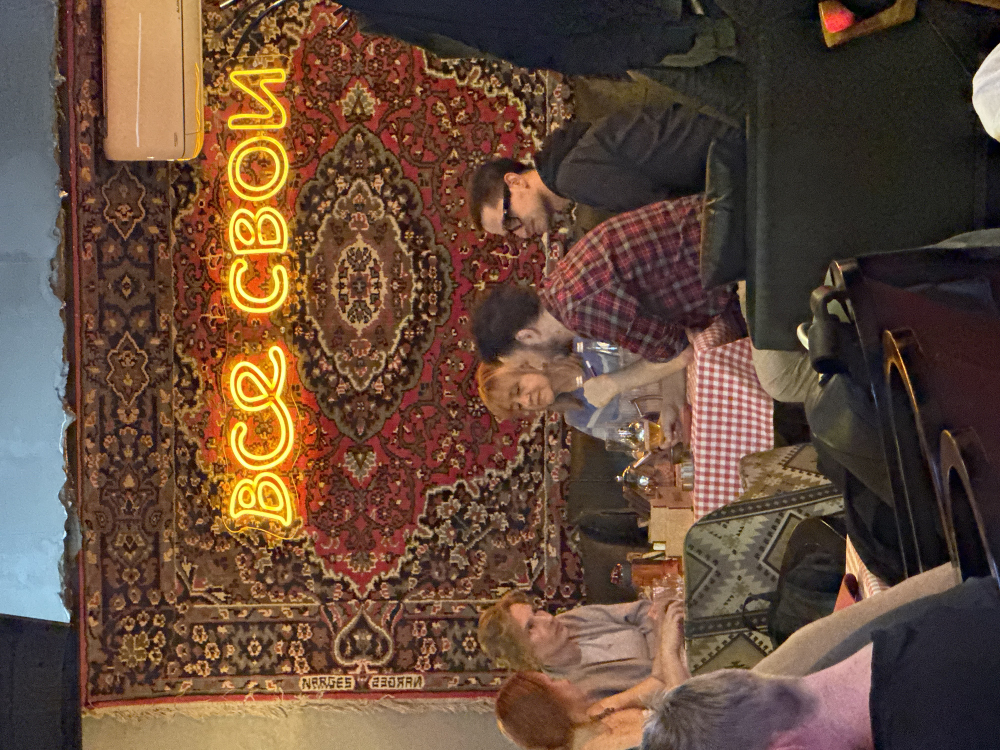 A scene in Tashkent, Uzbekistan, features a group of people at a table in a restaurant, with a large, patterned carpet covering a wall. An illuminated neon sign hangs near the carpet. The people around the table are engaged in conversation and appear to be enjoying themselves. The table is covered with a red-and-white checkered tablecloth.