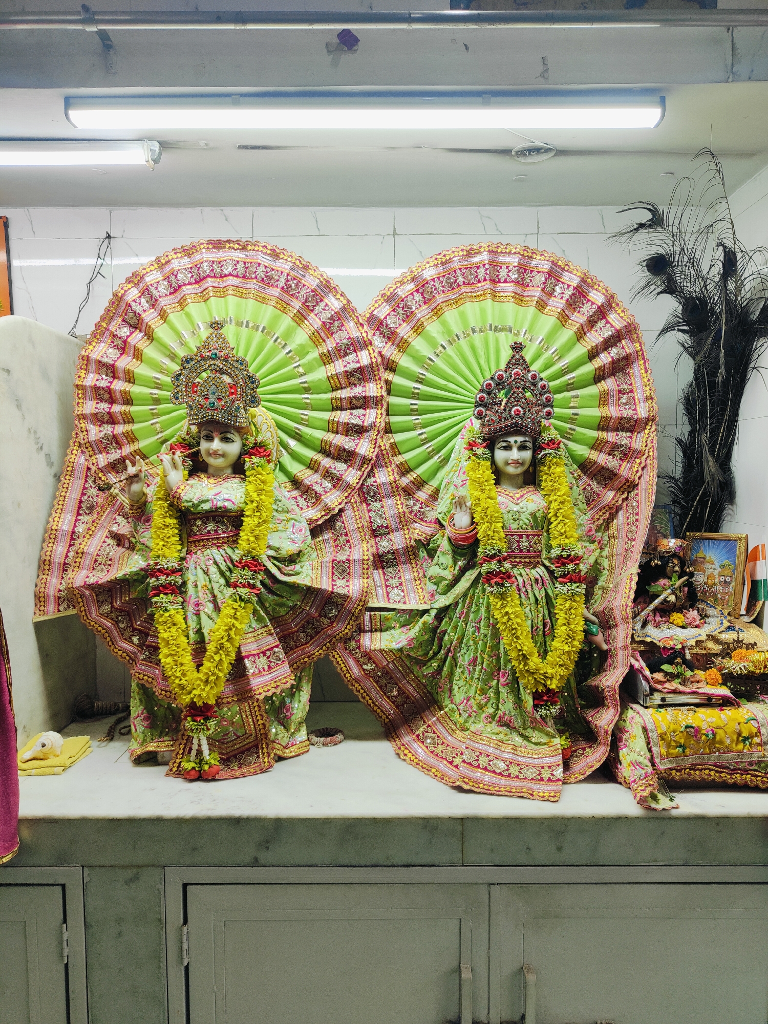 The image depicts two ornate statues of Hindu deities, likely Radha and Krishna, adorned in vibrant green and pink attire, elaborate crowns, and yellow floral garlands. They are positioned on a marble altar within a brightly lit interior space. Behind them, large, fan-like decorations in shades of green and gold fan out, intricately embroidered with metallic threads and red trim. To the right of the statues, a collection of smaller devotional items is visible, including a framed picture, an Indian flag, and floral offerings, set against a backdrop of peacock feathers. The statues are situated on a white marble platform, with what appear to be wooden cabinets below. The environment suggests a temple or shrine, possibly located in Gurgaon, India, given the presence of Hindu deities and Indian cultural elements. The lighting is bright and even, emanating from overhead fluorescent fixtures.
