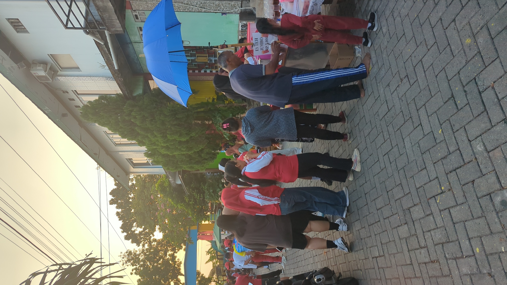 A group of people are gathered on a paved walkway in front of a building that appears to be a commercial establishment or community center, with trees and a bright sky in the background. A blue umbrella is open, sheltering a food or drink stall. Several individuals are standing in line at the stall, which displays a sign that reads "ES TEH JUMBO RP 5.000". Some people are wearing red clothing, suggesting a possible event or uniform. The environment appears to be an outdoor public space. Based on the context of the sign and the general ambiance, this scene is likely in Semarang, Indonesia.