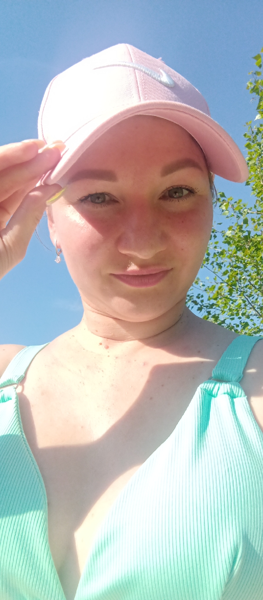 A woman is centered in a low-angle, sunlit selfie, wearing a light pink baseball cap and a light green ribbed swimsuit. She is fair-skinned with light brown hair and eyes and is holding the brim of the cap with her right hand. She has a necklace and small earrings. The background is a clear blue sky with some green tree foliage visible in the upper right corner. The location is Unknown, Unknown.