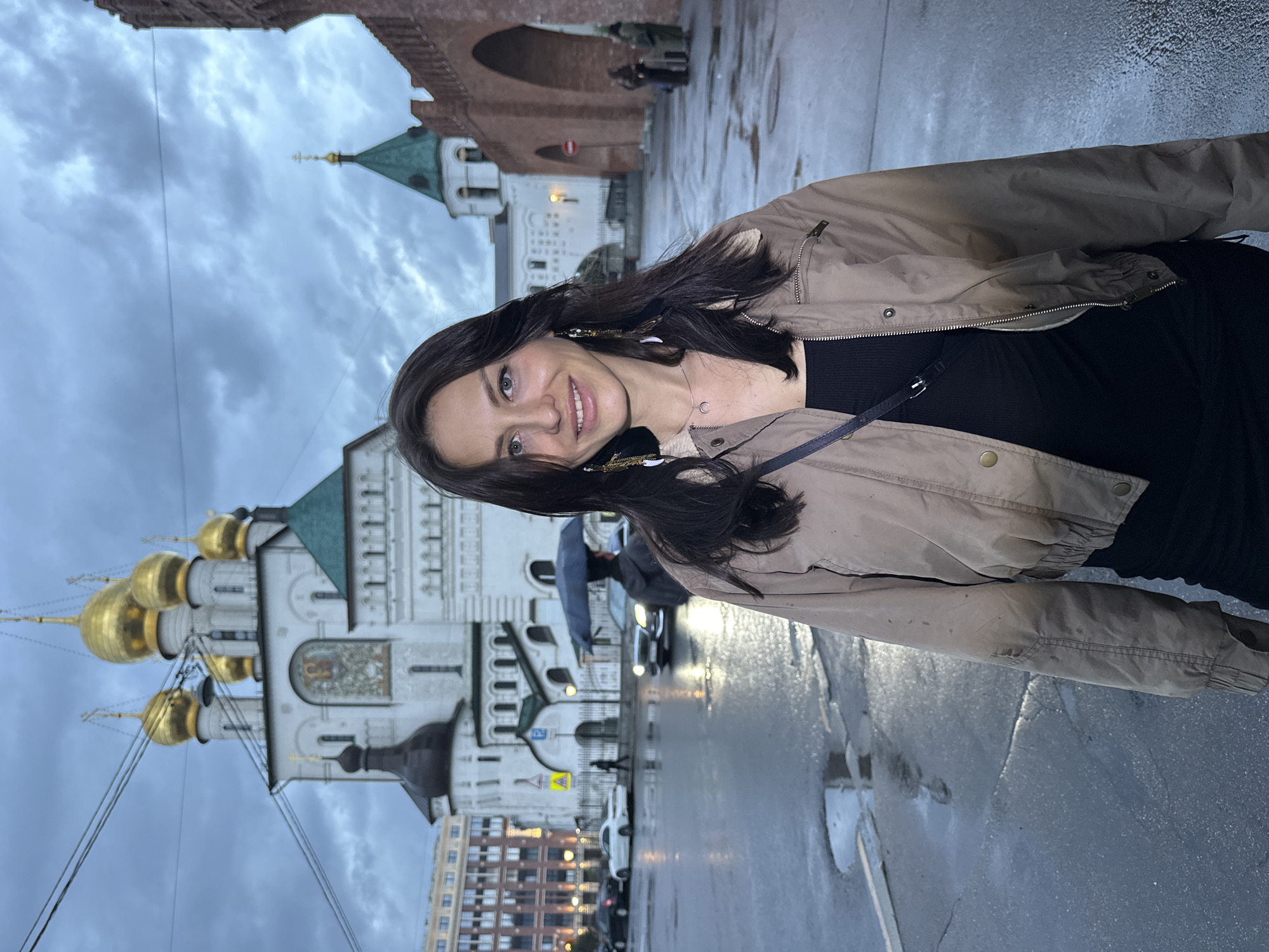 A medium shot, eye-level, outdoor, slightly overcast, full-body, portrait photograph features a woman posing in the foreground. The woman has long, dark brown hair and is smiling at the camera. She is wearing a tan bomber jacket over a black ribbed dress and a silver necklace with a crescent moon pendant. She also has long, dark earrings with gold and white accents. The background depicts a street scene in front of a white Orthodox church with golden domes, and a red brick wall with arches. The street is wet, indicating recent rain, with puddles reflecting the cloudy sky. Several cars are visible on the street, and a person is holding an umbrella. The overall impression is of a casual moment captured in a historic urban setting. The location is identified as the Moscow Kremlin, in Moscow, Russia.