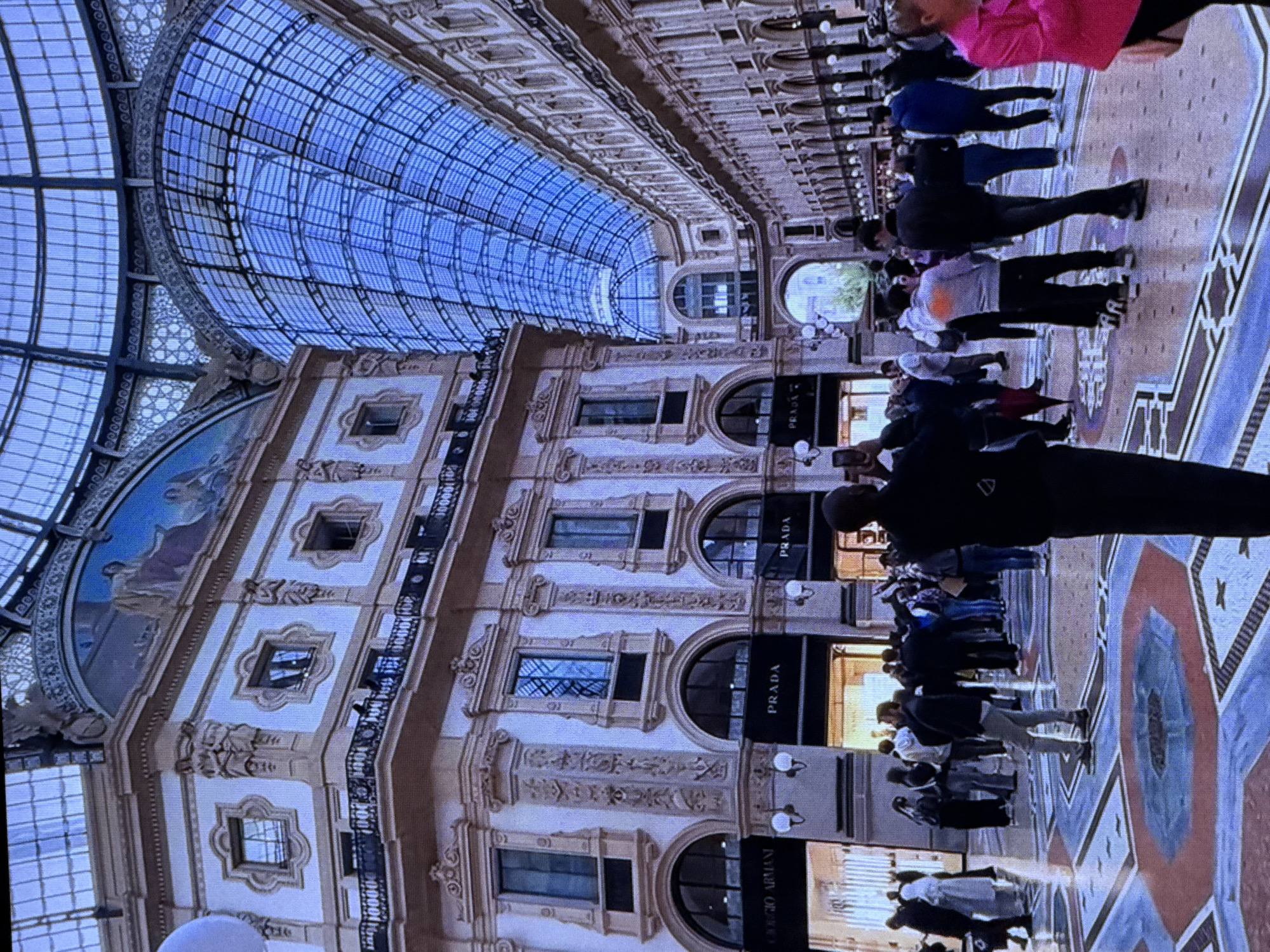 The image depicts the interior of a grand, domed shopping arcade with a blue, glass-paneled ceiling. The architecture is ornate, featuring classical columns, arched entrances, and decorative facades. Several high-end retail stores, including "Giorgio Armani" and "Prada," are visible along the sides. A diverse crowd of people is present, some walking, some standing, and some taking photographs. The floor is a mosaic pattern of various colors and geometric shapes. The scene suggests a public space, likely a popular tourist destination, characterized by its opulent design and busy atmosphere. The specific location is the Galleria Vittorio Emanuele II in Milan, Italy, not Dubai, United Arab Emirates.