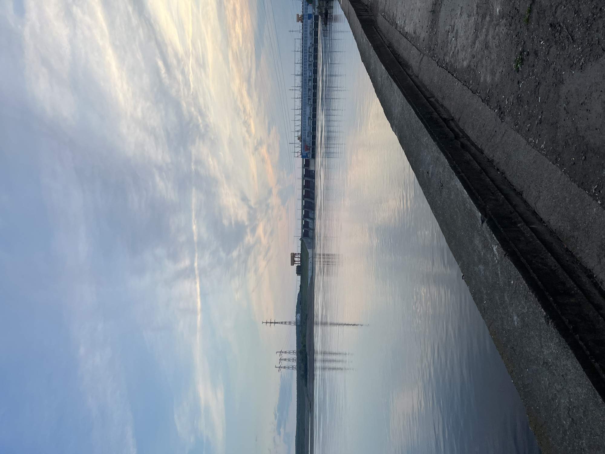 The image captures a vast, serene landscape featuring a large body of water, likely a river or reservoir, under a soft, diffused sky. In the foreground, a concrete embankment or pathway runs along the right edge, providing a sharp textural contrast to the smooth water surface. The water is remarkably calm and reflective, mirroring the gentle hues of the sky and the distant structures.
On the far bank across the water, a significant hydroelectric power plant or dam complex stretches horizontally. This industrial structure consists of multiple sections, including what appears to be a power station building and various control gates, with numerous power lines extending from it. Further along the distant bank to the left, several large electricity transmission towers (pylons) are visible, their angular forms casting subtle reflections onto the water.
The sky above is partly cloudy, displaying a blend of light blue, soft grey, and delicate hints of warm pink or orange, characteristic of either sunrise or sunset. The overall lighting is soft and even, contributing to the tranquil atmosphere. No people or active events are depicted, making the scene a static portrayal of a landscape where natural elements meet human-made infrastructure. There is no visible text in the image. Given the location context of Chaikovsky, Russia, this scene likely depicts the Kama River and its associated hydroelectric facilities.