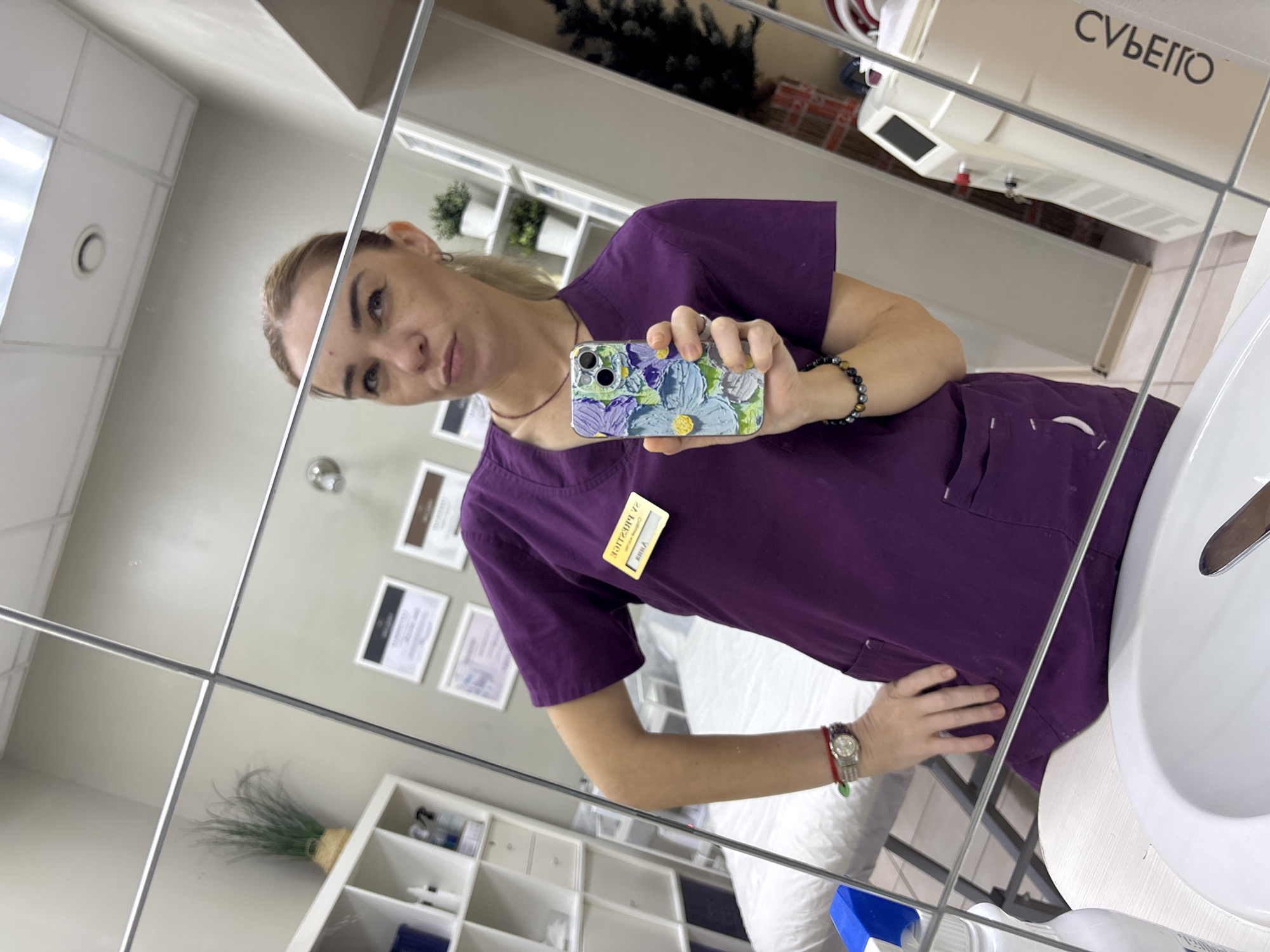A blonde woman in a purple scrub top and holding a floral phone takes a mirror selfie in a room that appears to be a medical or aesthetic clinic. The woman has her left hand on her hip and is looking towards the camera. A name tag on her scrub top reads "Anna". The room contains shelves with various items, a treatment bed, and medical equipment. A large mirror occupies most of the image, reflecting the scene. The woman is wearing a watch and bracelets. There is a sink and a white countertop in the foreground. The overall environment suggests a professional setting. The city of Medovoye, Russia is not depicted in the image.