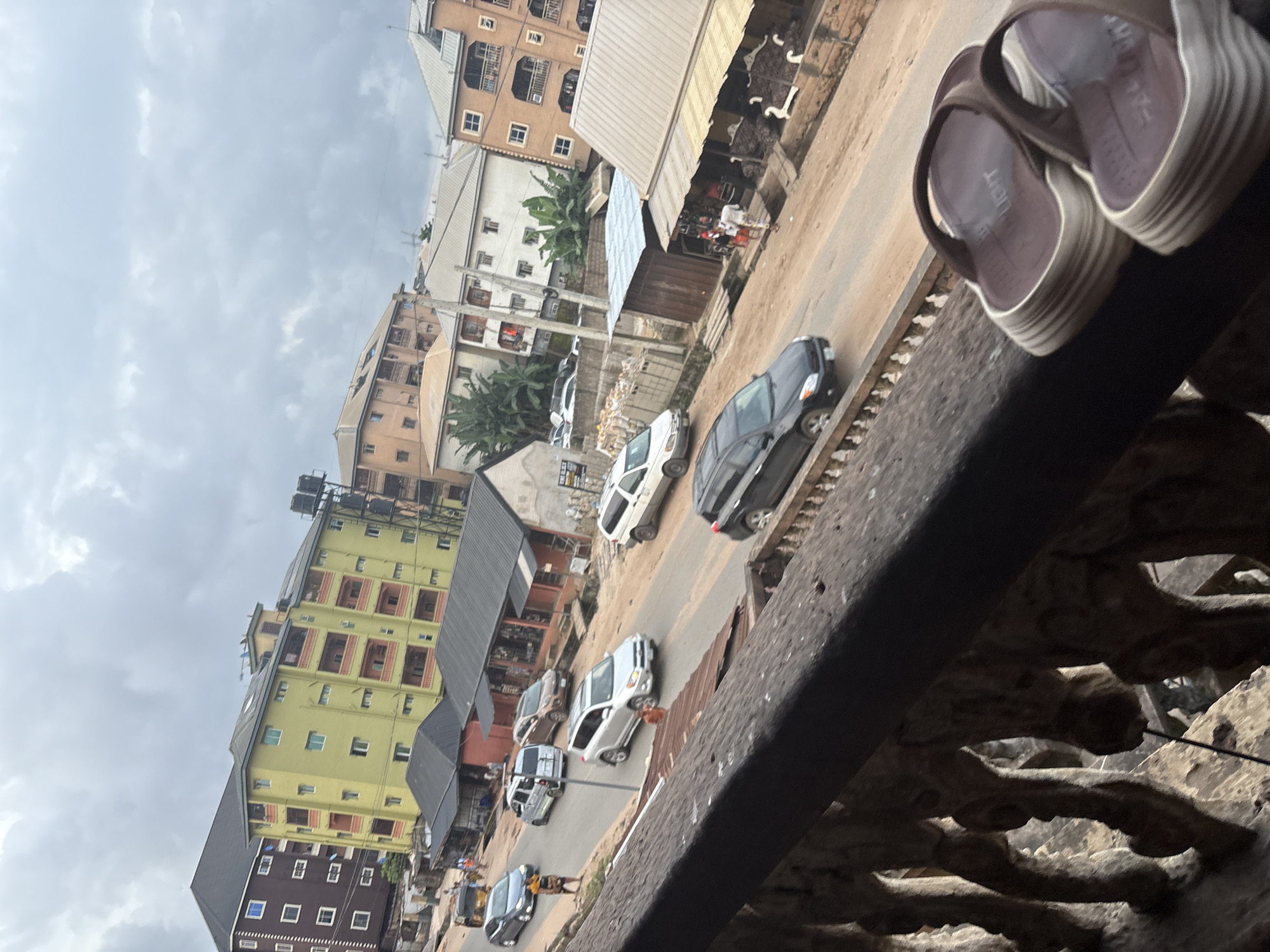A medium-angle, slightly tilted outdoor shot captures a street scene in Nkpor, Nigeria, on an overcast day. In the foreground, a dark brown wooden railing with carved vertical supports partially obscures the view. Resting on the railing are a pair of light brown and white slip-on sandals. The street below is paved with asphalt and shows signs of wear. Several vehicles are present: a silver SUV, a dark gray sedan, a silver minivan, and a white van. People are visible on the street, including a woman in a yellow headwrap and patterned dress walking on the sidewalk, and a few individuals near roadside stalls. The surrounding environment consists of multi-story buildings in various states of construction and completion, with different colored facades and visible windows. Some buildings have balconies, and one large yellow building has a water tank on its roof. Roadside stalls and makeshift structures are also present, suggesting a commercial area. The sky is heavily clouded, casting a diffused light over the scene.