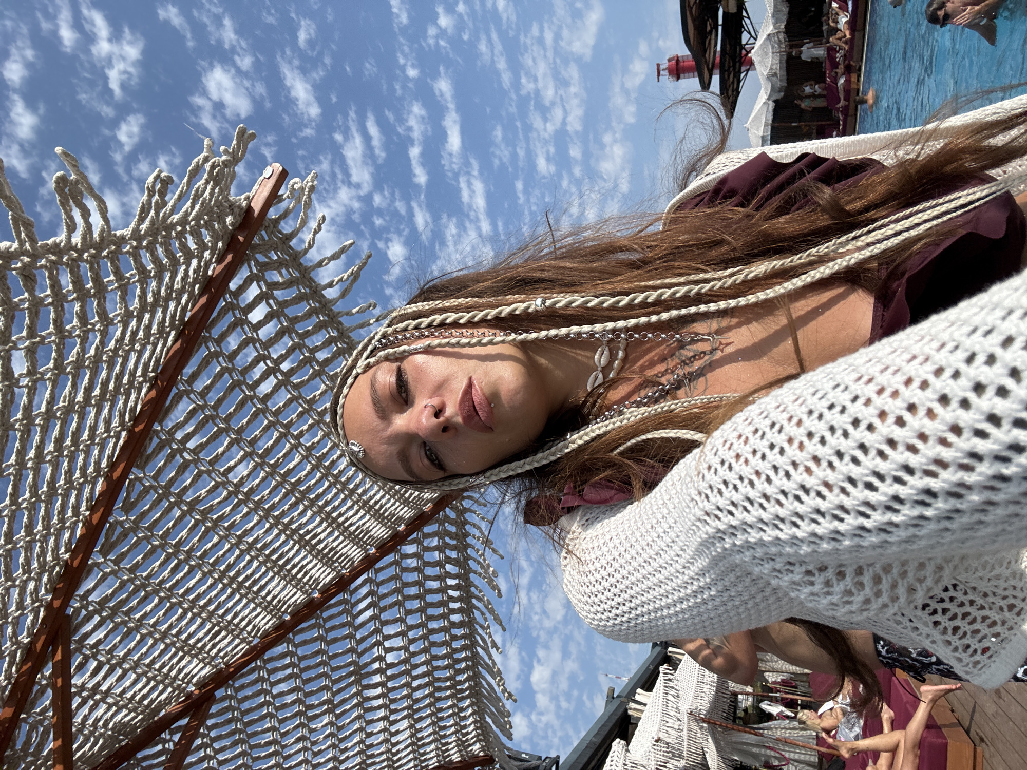 An overhead shot shows a woman with long brown hair styled in braids and adorned with a metallic headband. She is wearing a white, crocheted garment over a dark purple top and a shell necklace. Her gaze is directed towards the camera. In the background, several people are relaxing on lounge chairs near a swimming pool. The scene appears to be an outdoor venue, possibly a resort or beach club, under a blue sky with scattered clouds. The location is identified as Sochi, Russia.