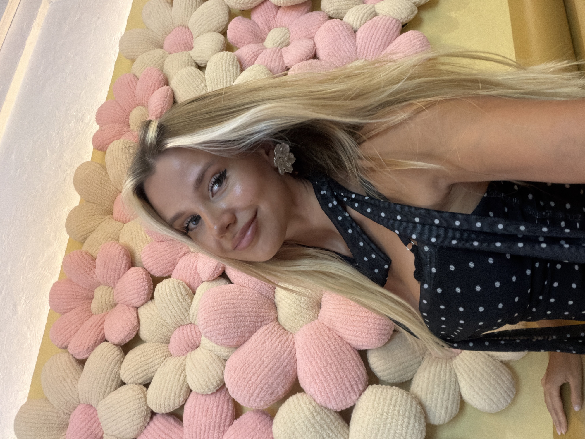 A young Caucasian woman with blonde hair and heavy eye makeup smiles into the camera. She is wearing a black polka dot dress and a silver flower earring. Behind her is a wall decorated with large, plush flowers in shades of pink and cream. The scene appears to be indoors, possibly in a cafe or boutique. The woman's left hand is resting on a surface to her lower left.