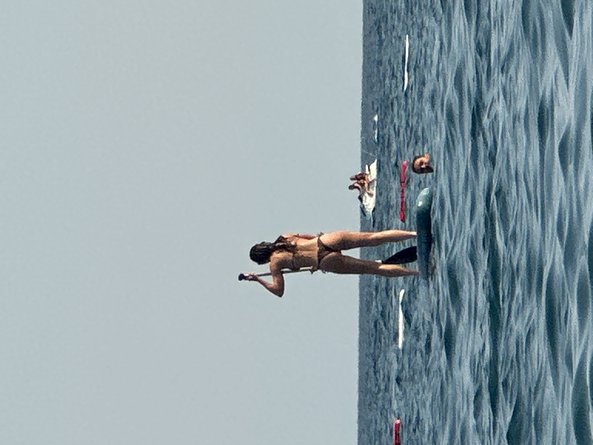In the city of Boreti, Montenegro, a woman in a brown bikini stands on a paddleboard in the ocean, holding a paddle. In the background, a jet ski carries two individuals. To the right of the paddleboarder, a person's head is visible above the water. The sea is calm with moderate waves, and the sky is clear and pale blue. Several other inflatable objects are scattered in the water.